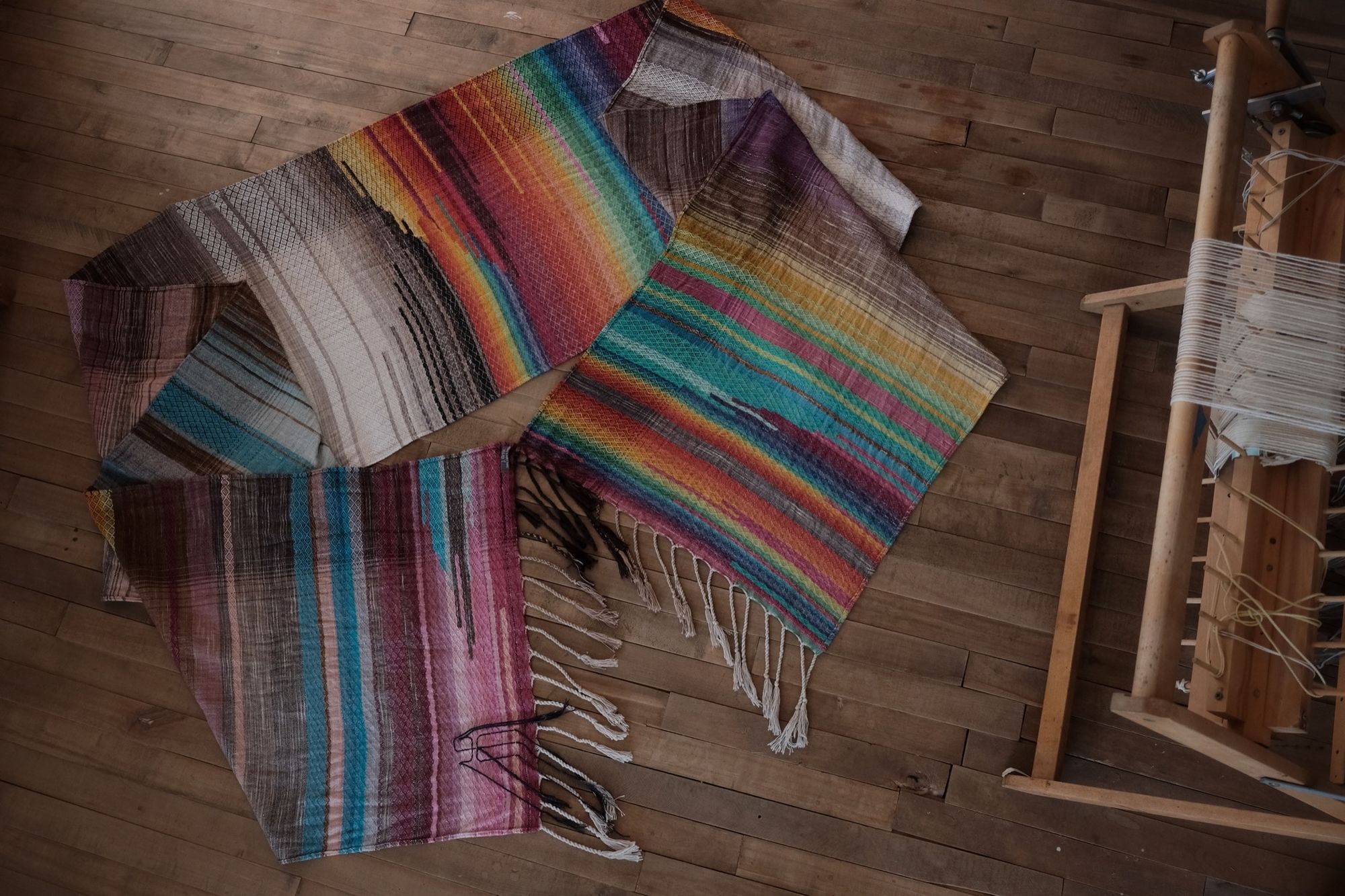 handwoven raw silk fabric woven with a diamond pattern in rainbow, gray, brown and cream colors on a wood floor with a loom in the corner