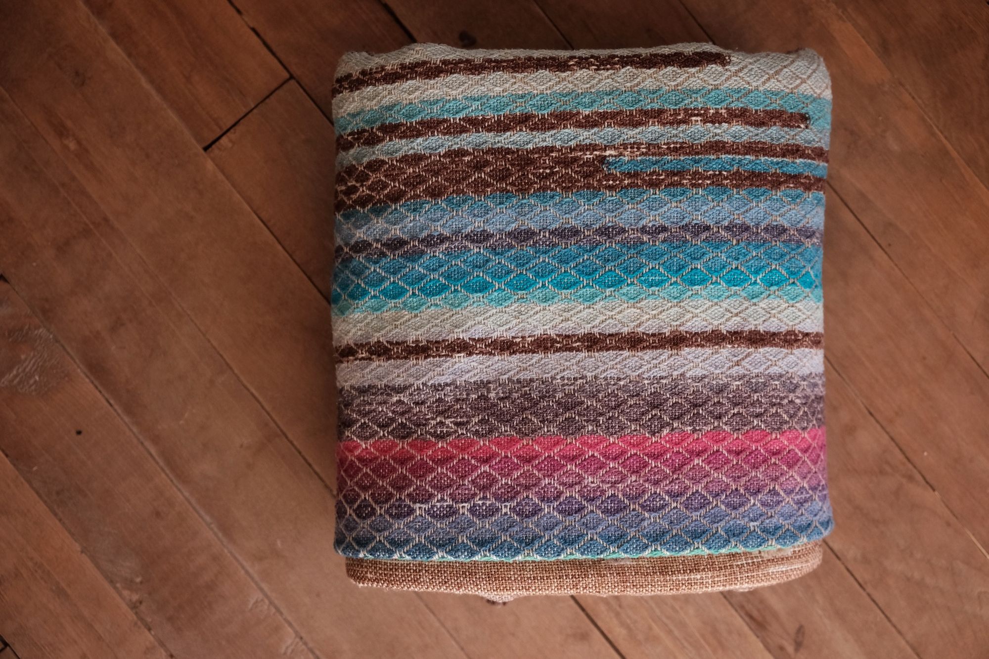 Handwoven fabric with a diamond weave pattern in shades of brown, blue and rainbow lays on a wooden floor