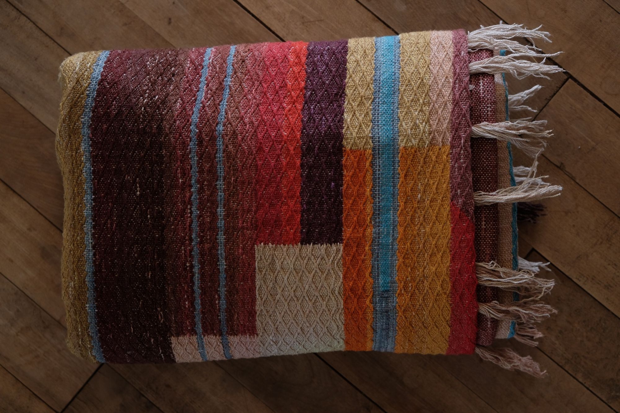 Handwoven fabric with a diamond texture pattern in natural browns, grey, purples, reds, turquoise, yellow, orange and pink. It is folded on a wooden floor.