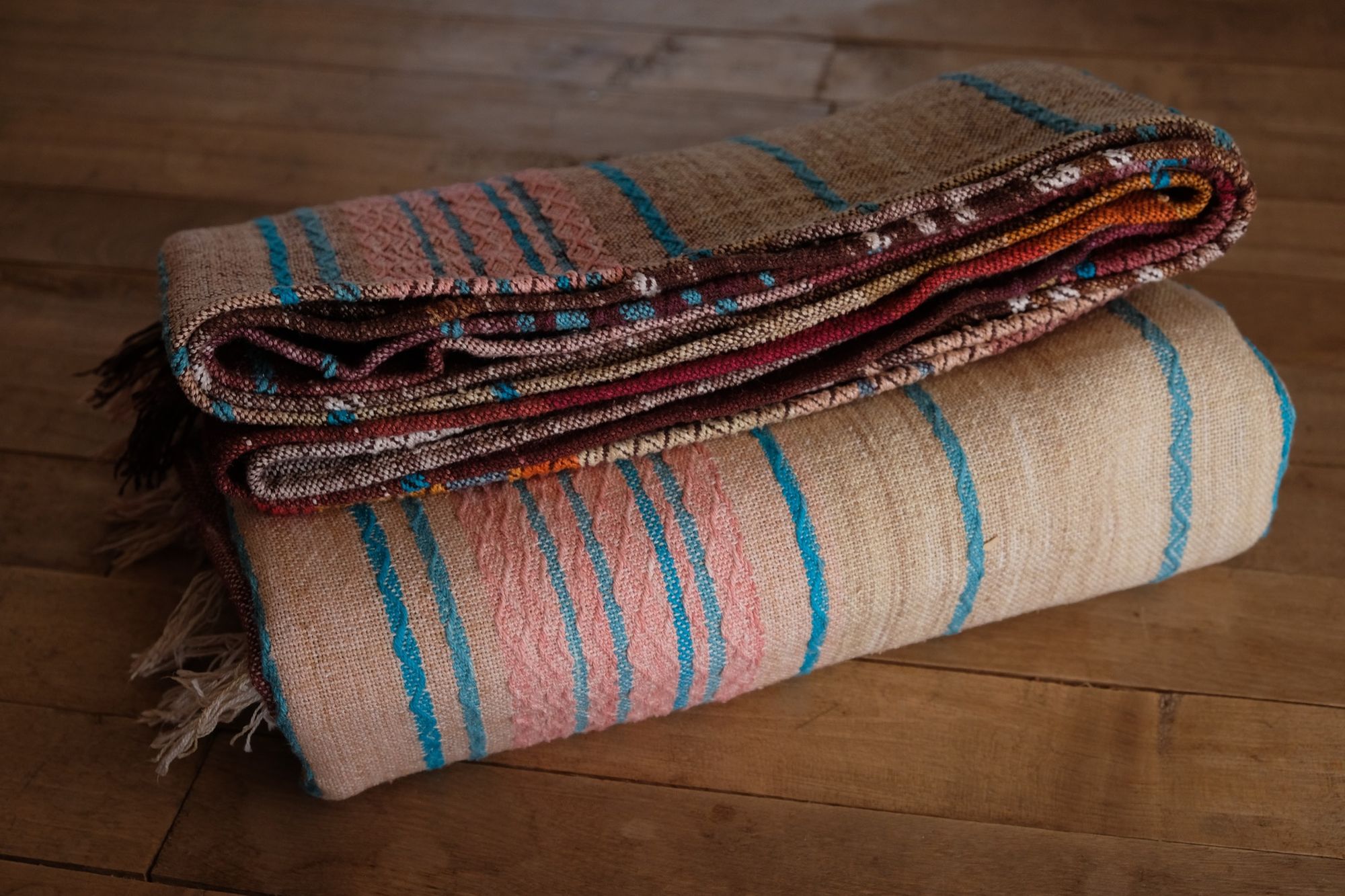 Handwoven fabric with a diamond texture pattern in natural browns, grey, purples, reds, turquoise, yellow, orange and pink. It is folded on a wooden floor.