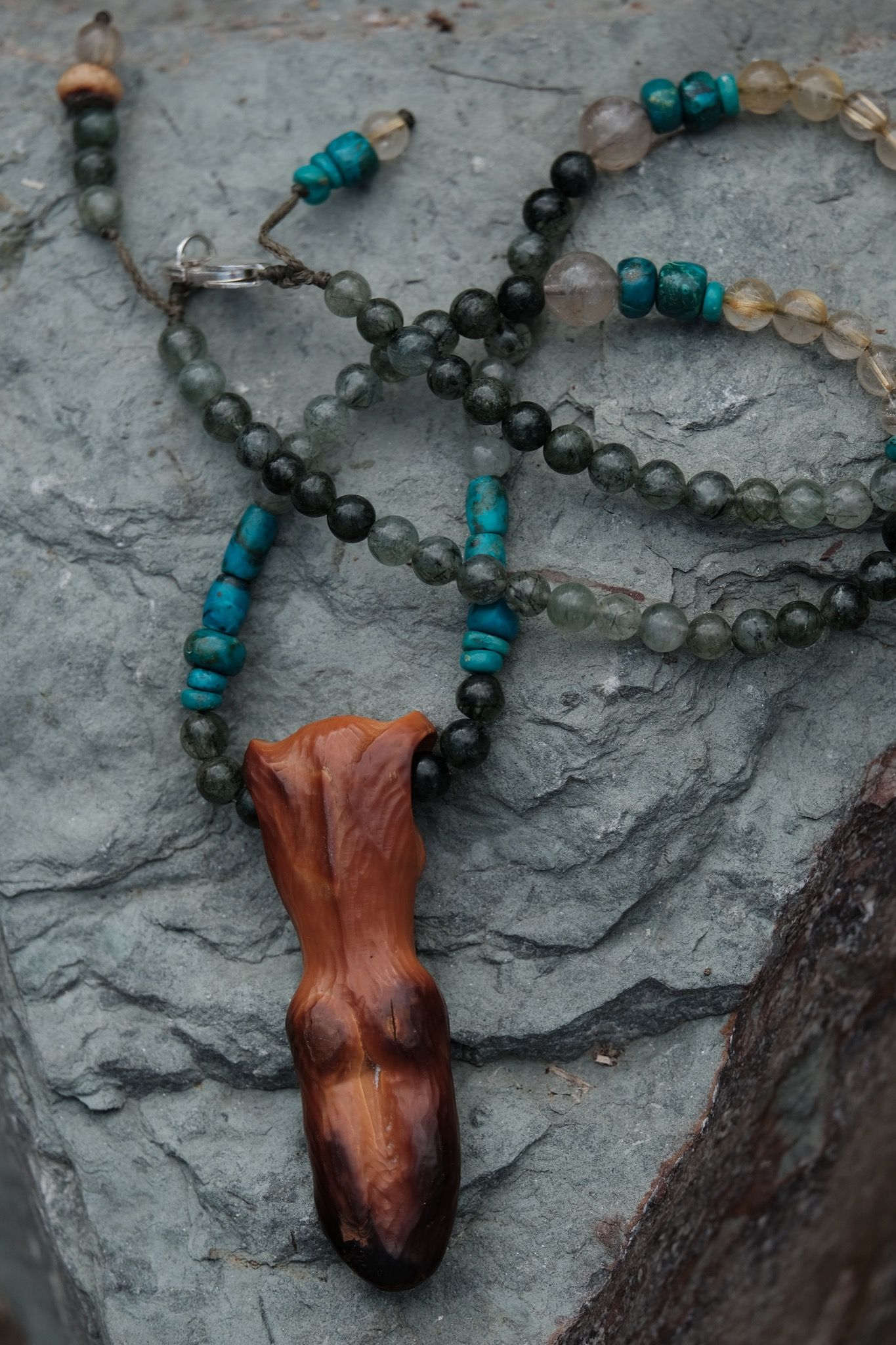 A necklace of green, blue, golden and Carmel ivory stones with a goddess figure at the center rests on a grey-blue stone