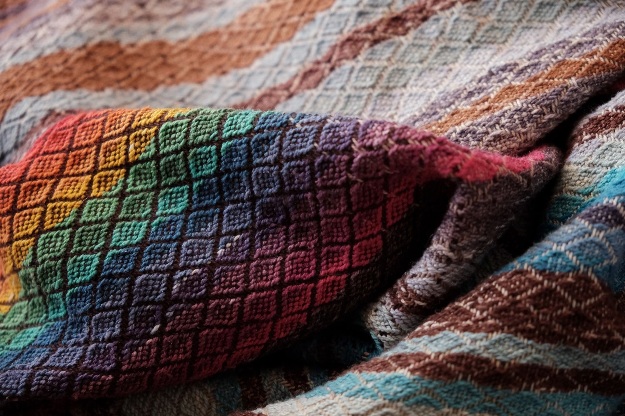 Handwoven fabric with a diamond weave pattern in shades of brown, blue and rainbow lays on a wooden floor