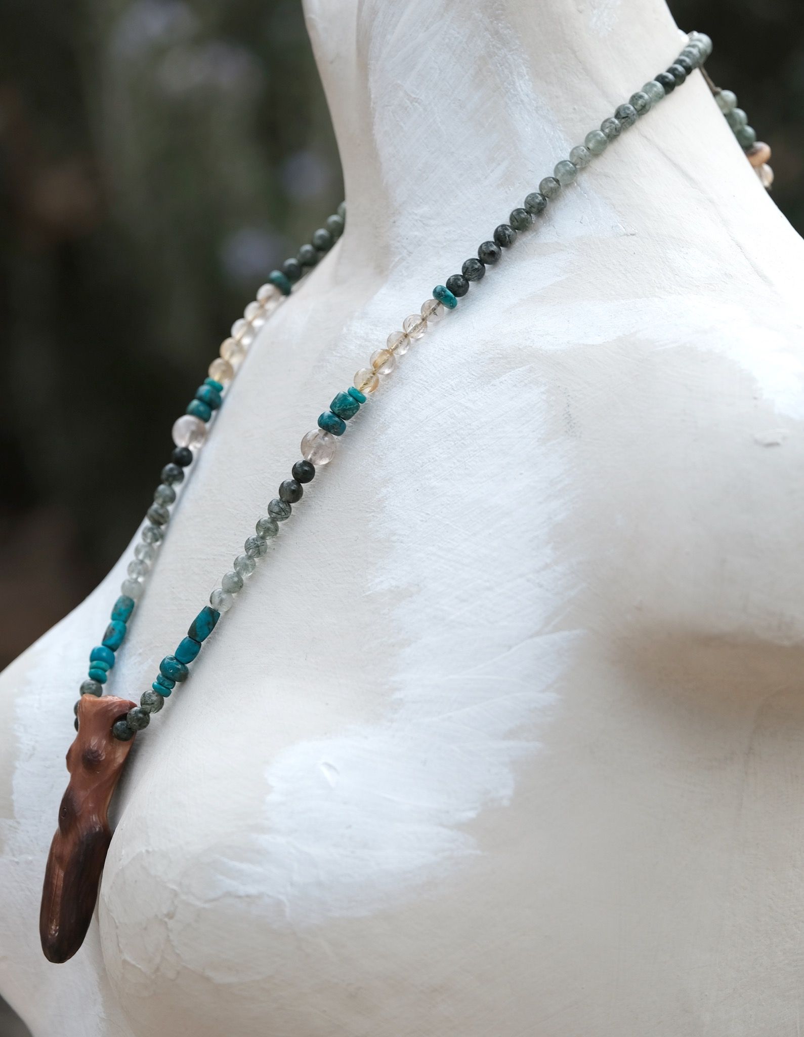 A necklace of green, blue, golden and Carmel ivory stones with a goddess figure at the center rests on a white painted mannequin