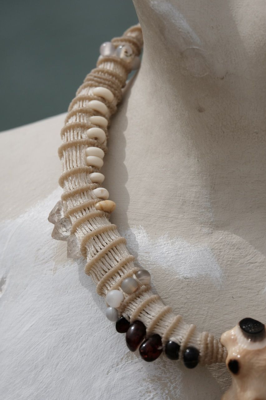 A white mannequin bust by the water wears a white sculptural tubular knot work necklace with beads and a walrus tooth goddess at the center