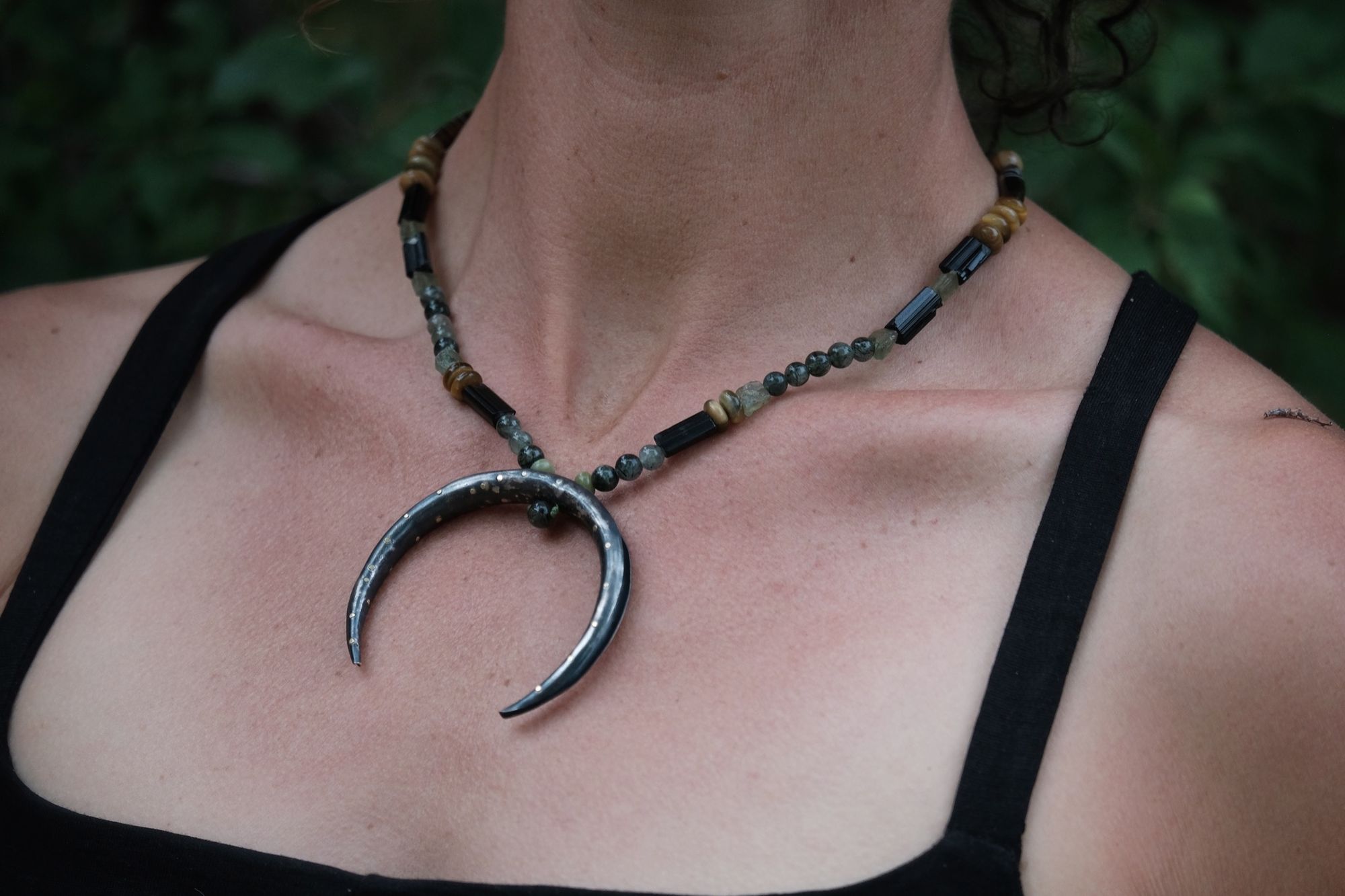 A necklace of brown, green, tan and black semi precious stone beads with a silver and gold moon at the center rests on a woman in a black dress
