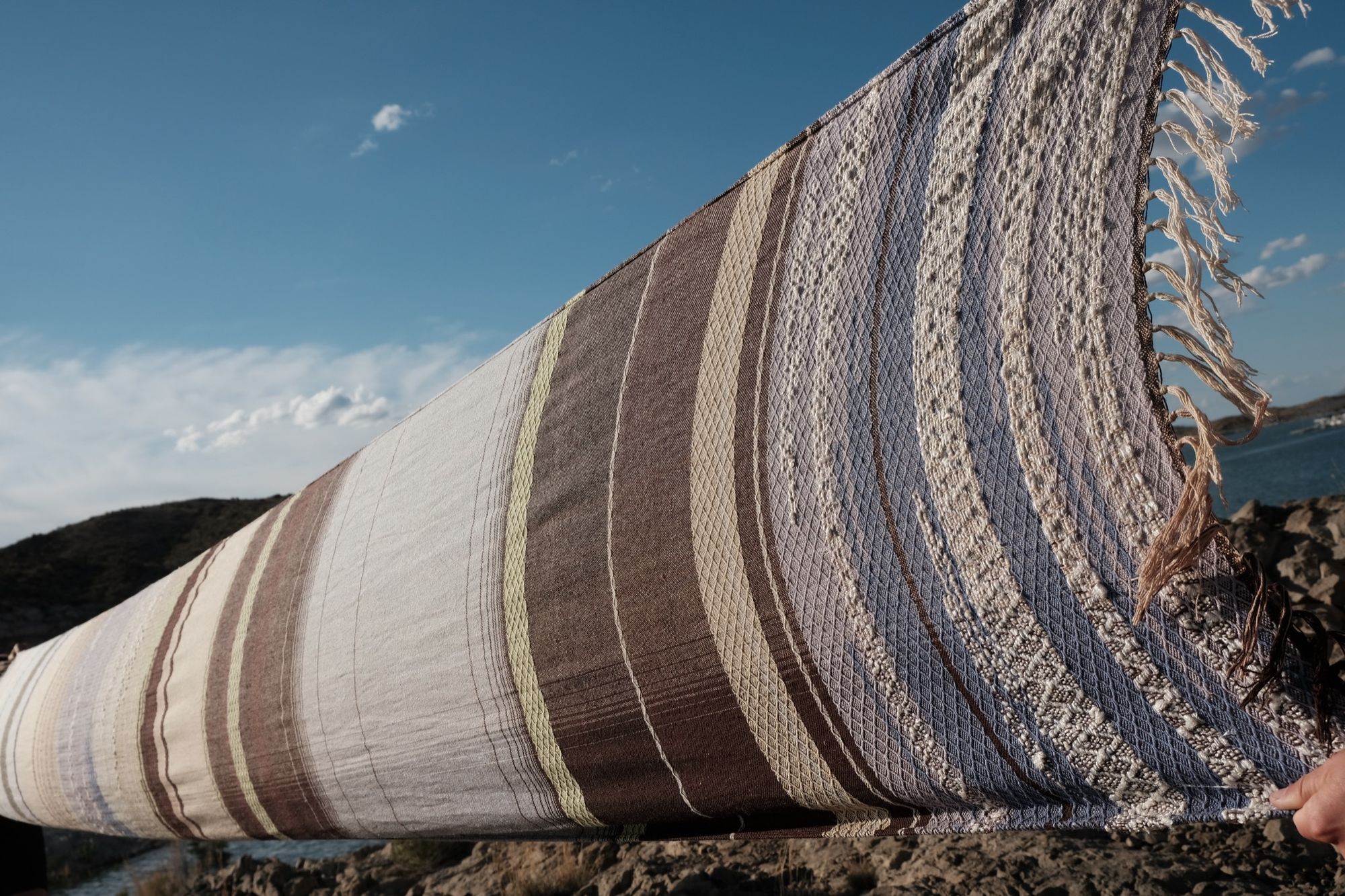 A long piece of handwoven fabric in lilac purple, dark brown and earth tones is stretched out against a blue sky