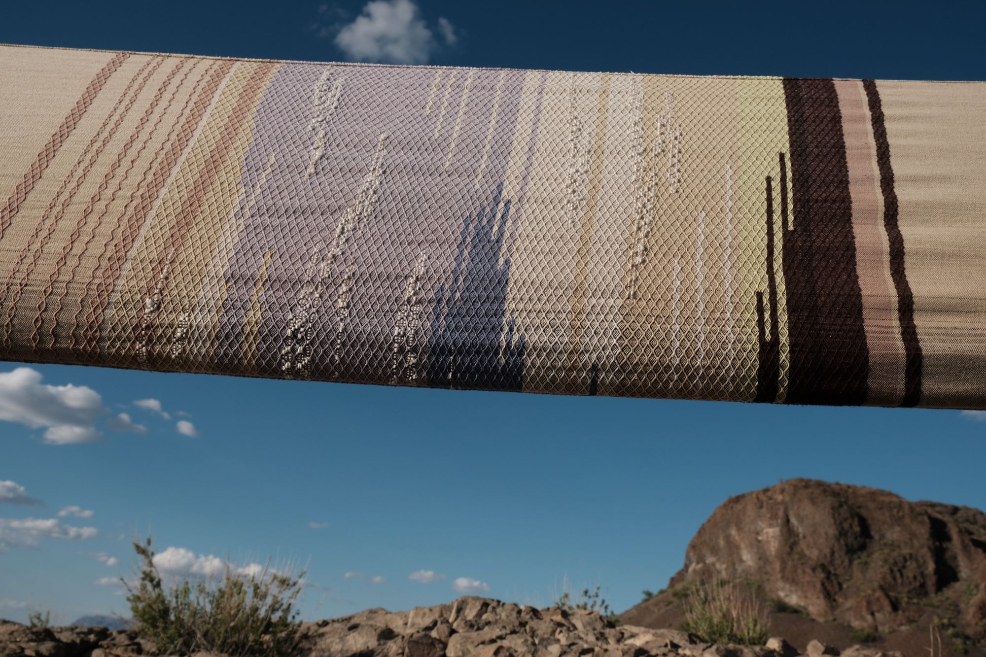 A long piece of handwoven fabric in lilac purple, dark brown and earth tones is stretched out against a blue sky