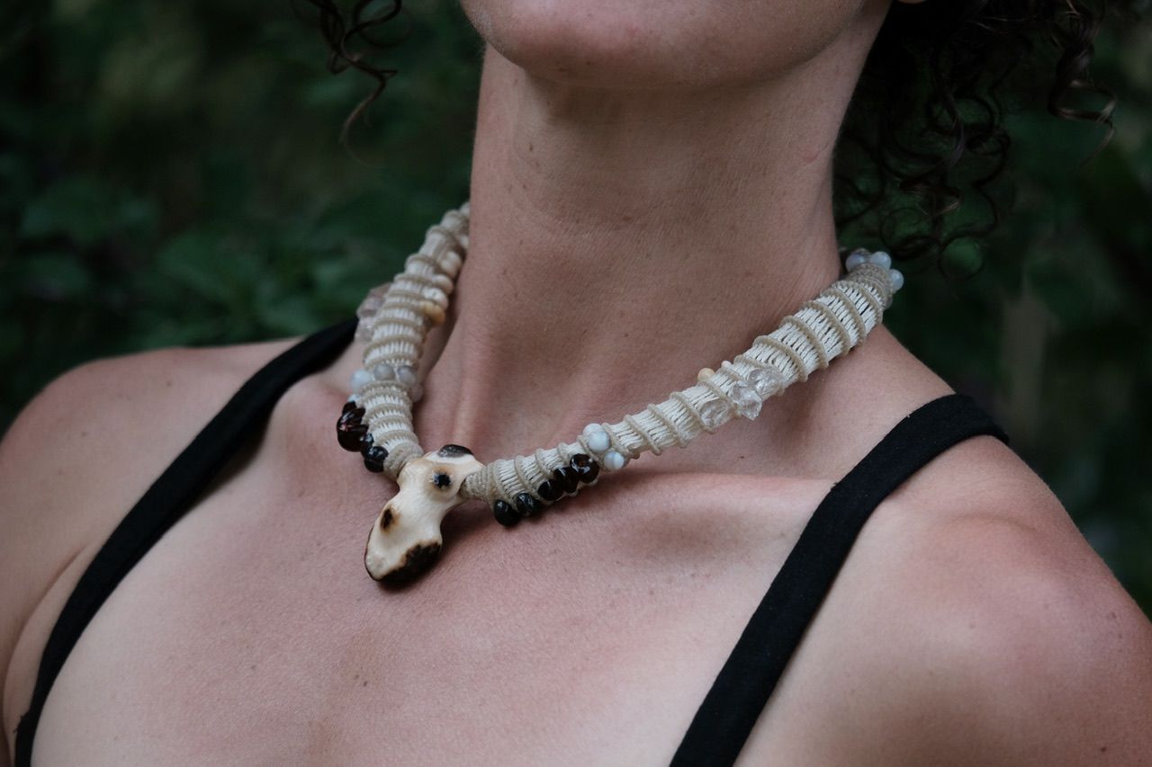 A woman in a black dress in the forest wears a white sculptural tubular knot work necklace with beads and a walrus tooth goddess at the center
