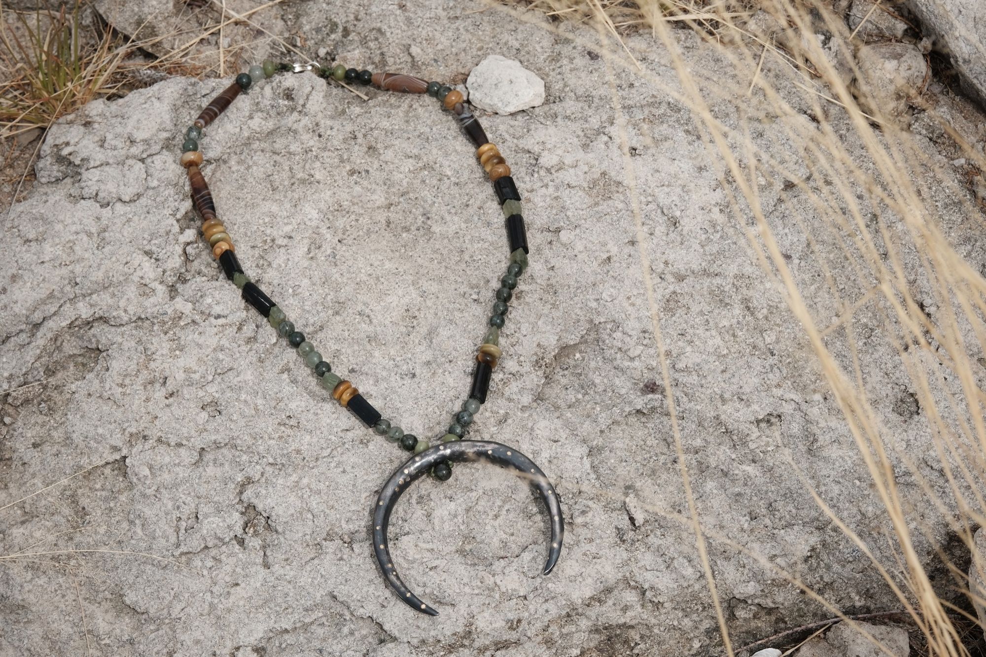 A necklace of brown, green, tan and black semi precious stone beads with a silver and gold moon at the center rests on white stones in the grass
