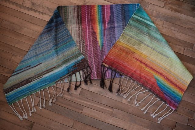 A handwoven diamond pattern rainbow shawl rests on a wood floor 
