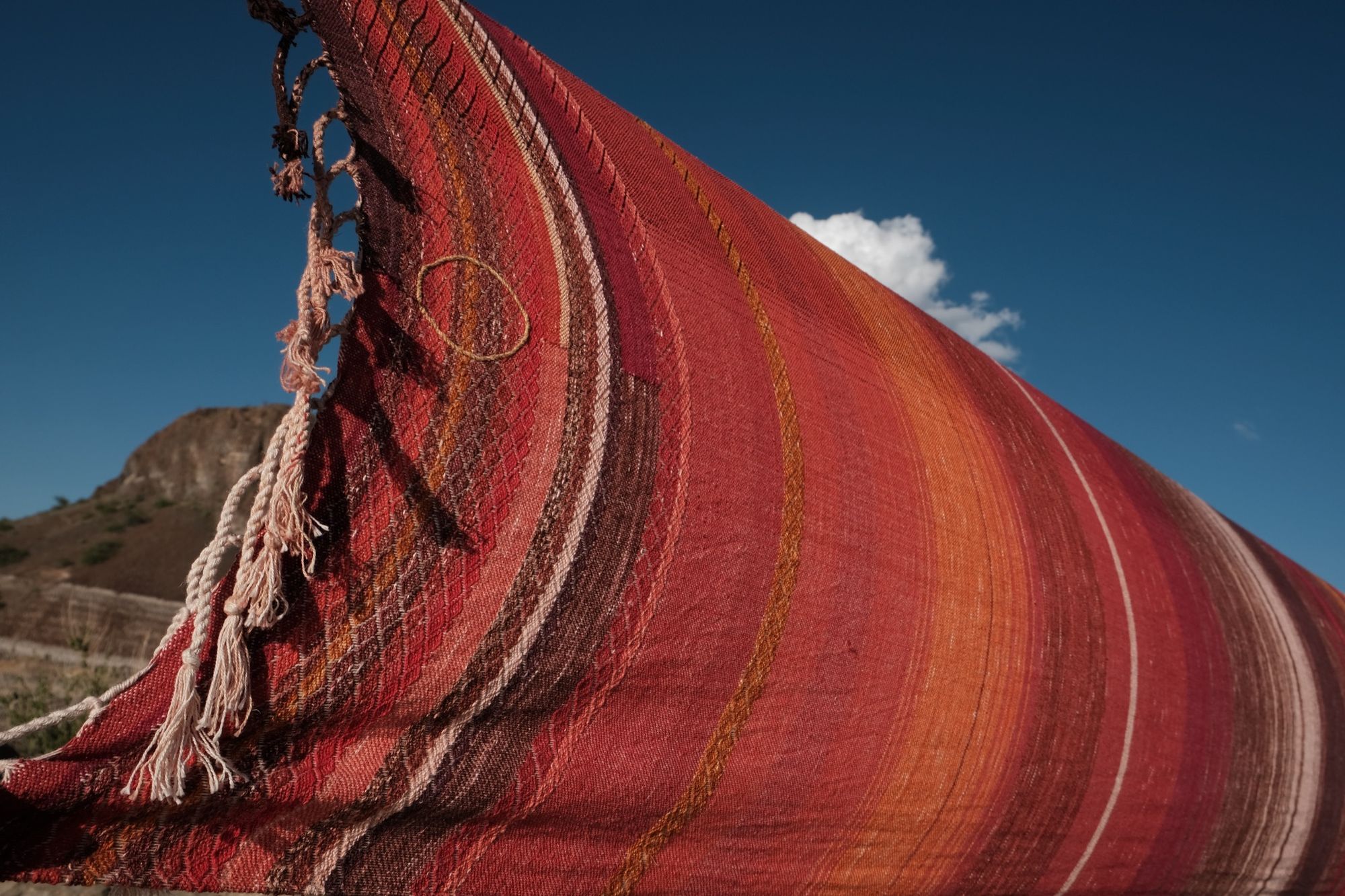 a long length of handwoven fabric in deep reds, purple, pink and orange is stretched across a blue sky with Rocky Mountains behind