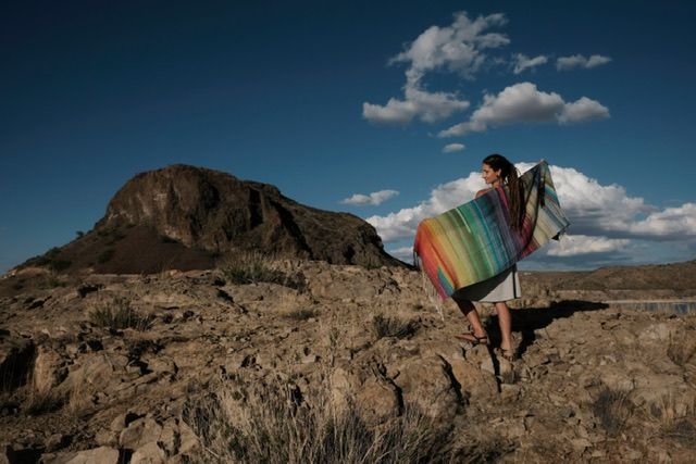 A woman wearing a handwoven many rainbow colored shawl stands in bright desert light on a Rocky Mountain With vivid blue sky and dramatic clouds behind