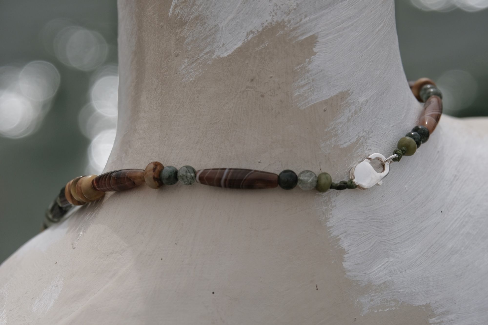 A necklace of brown, green, tan and black semi precious stone beads with a silver and gold moon at the center rests on a white mannequin