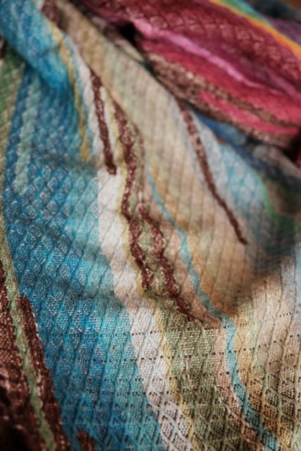 Detail of A handwoven diamond pattern rainbow shawl that rests on a wood floor 