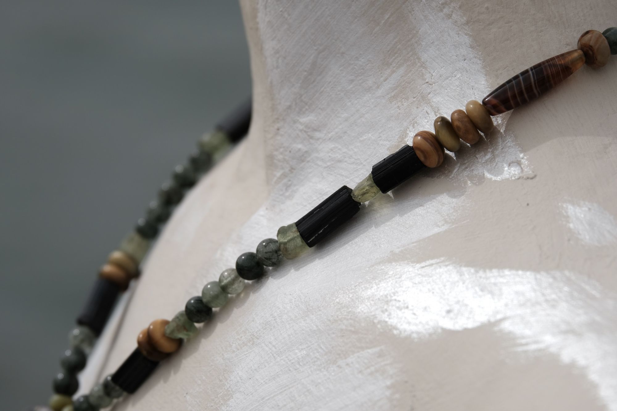A necklace of brown, green, tan and black semi precious stone beads with a silver and gold moon at the center rests on a white mannequin