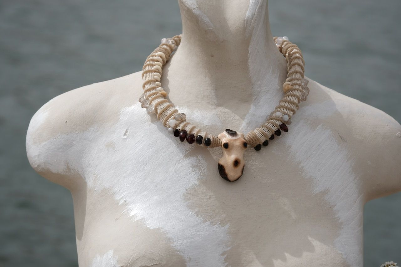 A white mannequin bust by the water wears a white sculptural tubular knot work necklace with beads and a walrus tooth goddess at the center