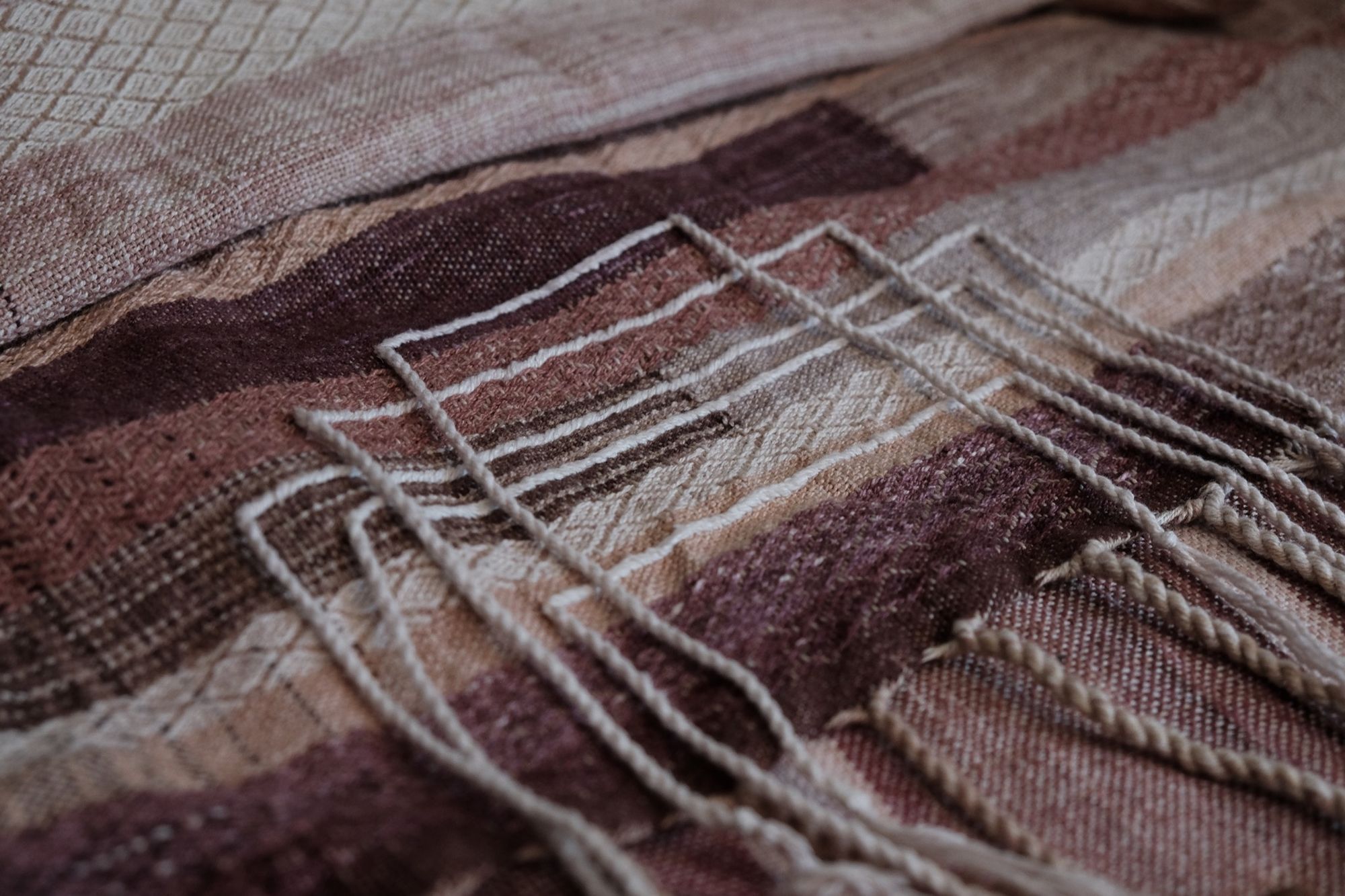 On a wooden floor rests handwoven fabric in shades of purple, mauve, burgundy and grey with geometric patterns, diamond weave and moon phases