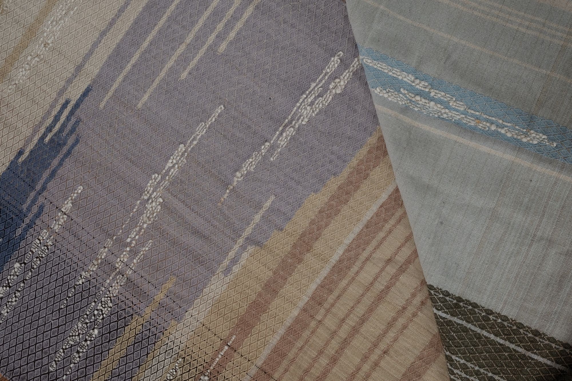 A long piece of handwoven fabric with diamond pattern in lilac purple, dark brown and earth tones is resting on a wooden floor