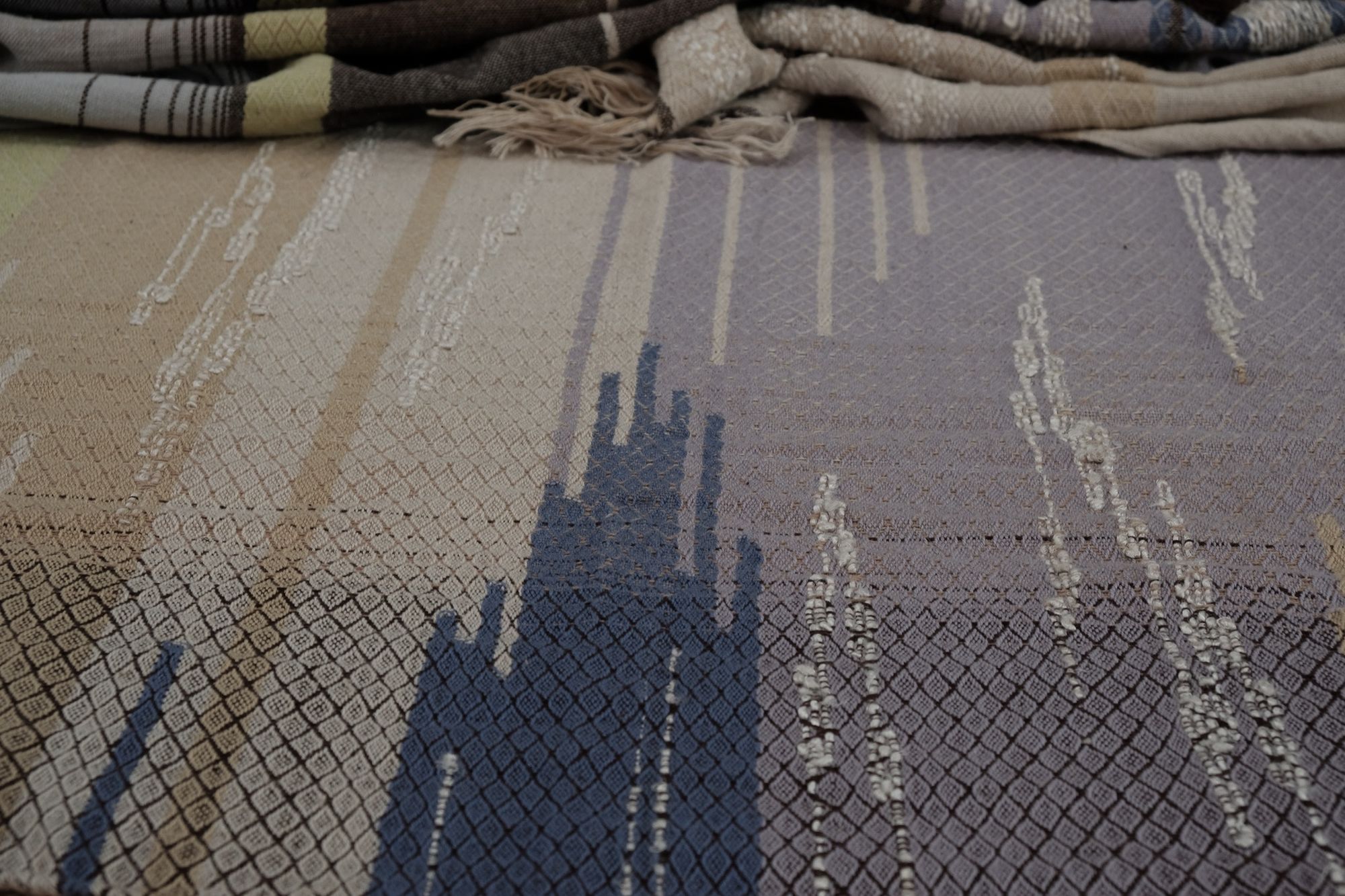 A long piece of handwoven fabric with diamond pattern in lilac purple, dark brown and earth tones is resting on a wooden floor