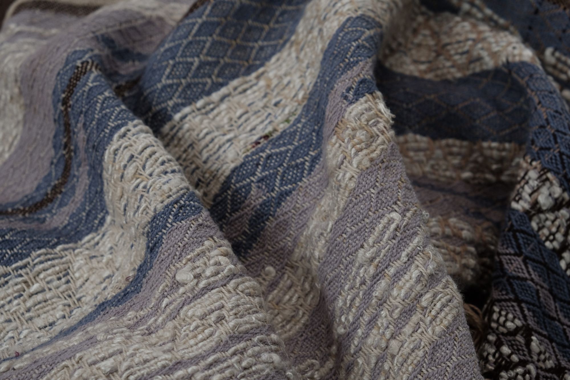A long piece of handwoven fabric with diamond pattern in lilac purple, dark brown and earth tones is resting on a wooden floor with details of banana silk shown prominently 