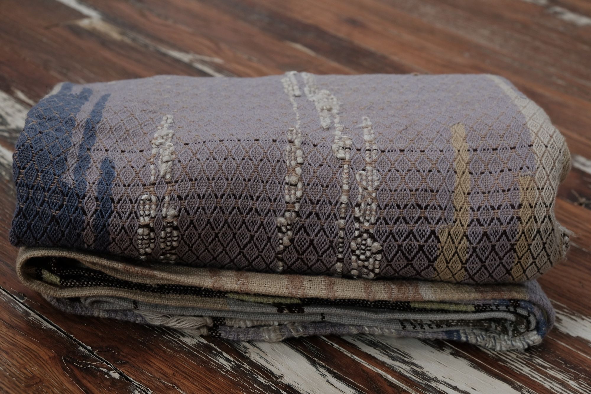A long piece of handwoven fabric with diamond pattern in lilac purple, dark brown and earth tones is folded on a wooden floor