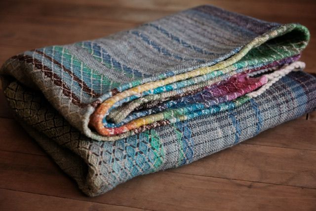 Detail of A handwoven diamond pattern rainbow shawl with fringe folded on a wood floor 