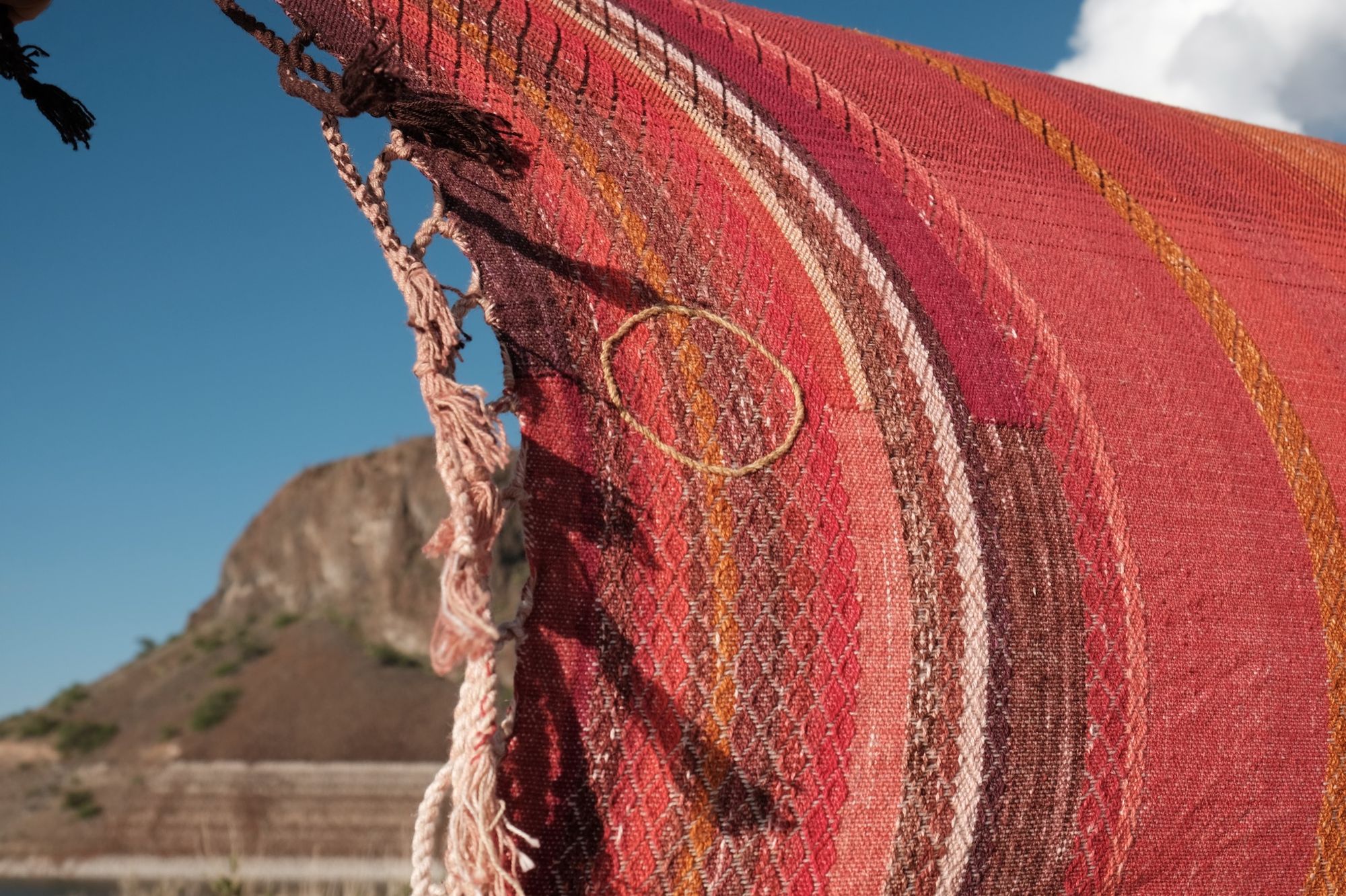 a long length of handwoven fabric in deep reds, purple, pink and orange is stretched across a blue sky with Rocky Mountains behind