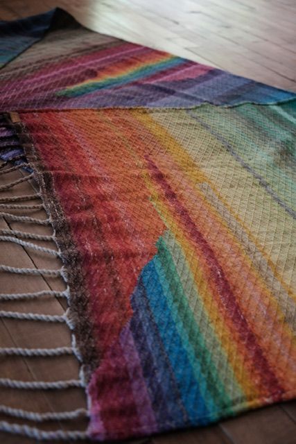 A handwoven diamond pattern rainbow shawl rests on a wood floor 