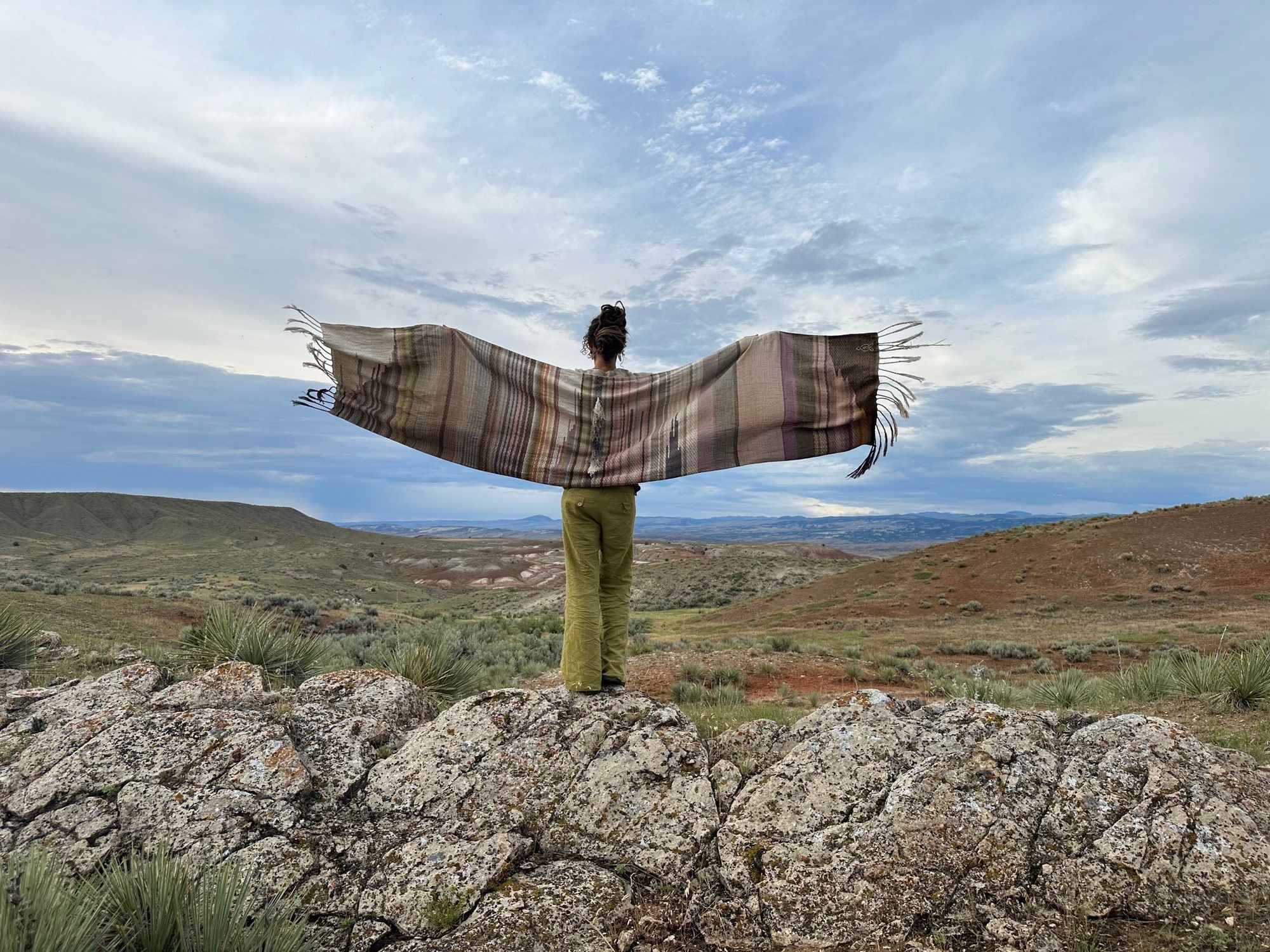 a woman wears a shawl of handwoven fabric of Naturally dyed subtle rainbow hues with a diamond texture pattern in a mountain landscape