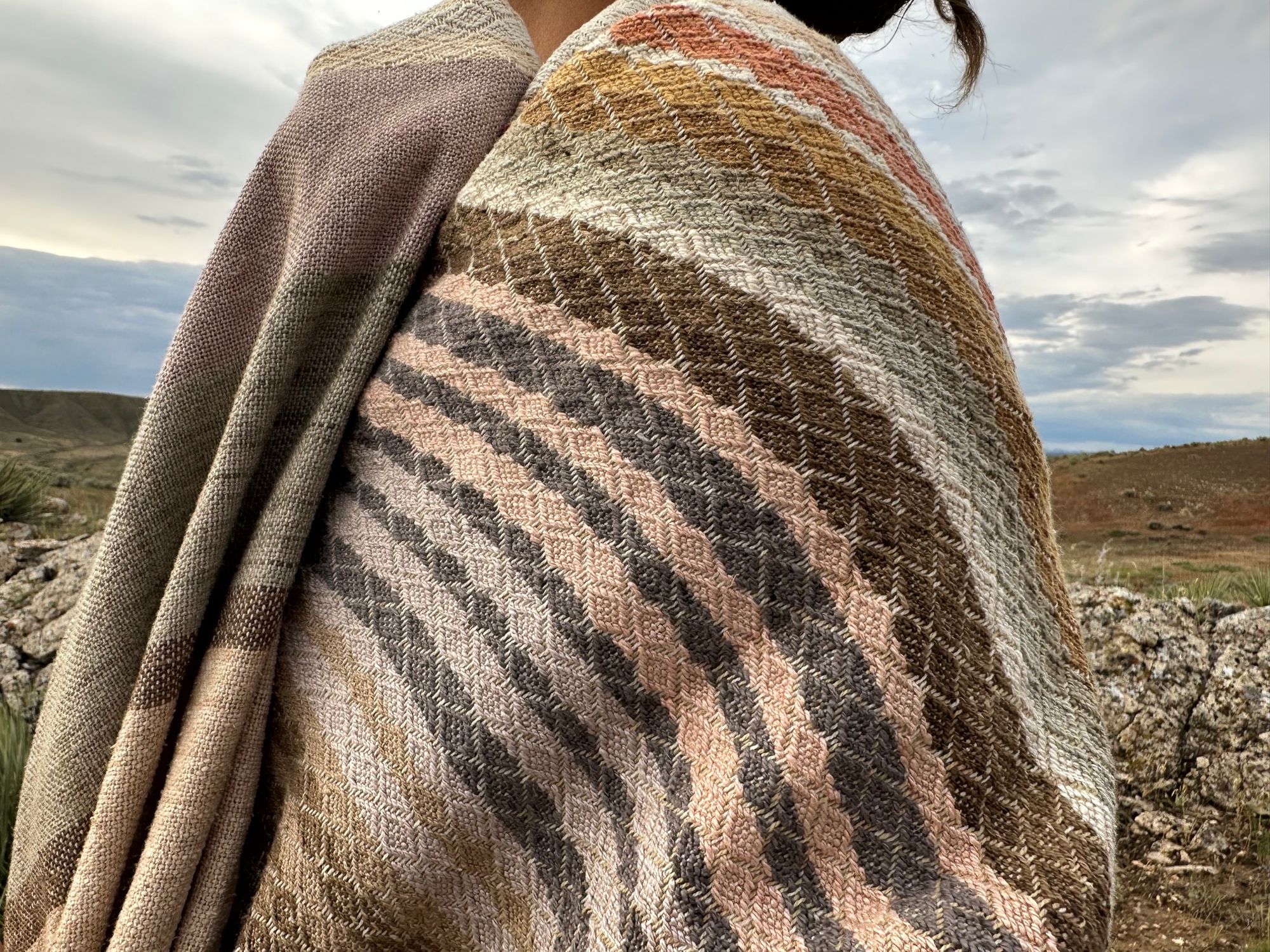 handwoven fabric of Naturally dyed subtle rainbow hues with a diamond texture pattern lays on a wooden floora woman wears a shawl of handwoven fabric of Naturally dyed subtle rainbow hues with a diamond texture pattern in a mountain landscape