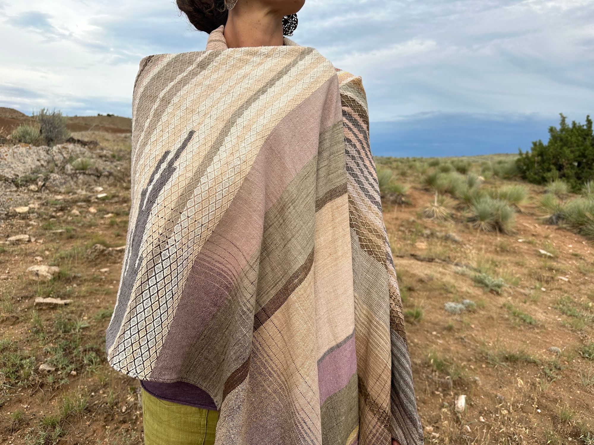 a woman wears a shawl of handwoven fabric of Naturally dyed subtle rainbow hues with a diamond texture pattern in a mountain landscape
