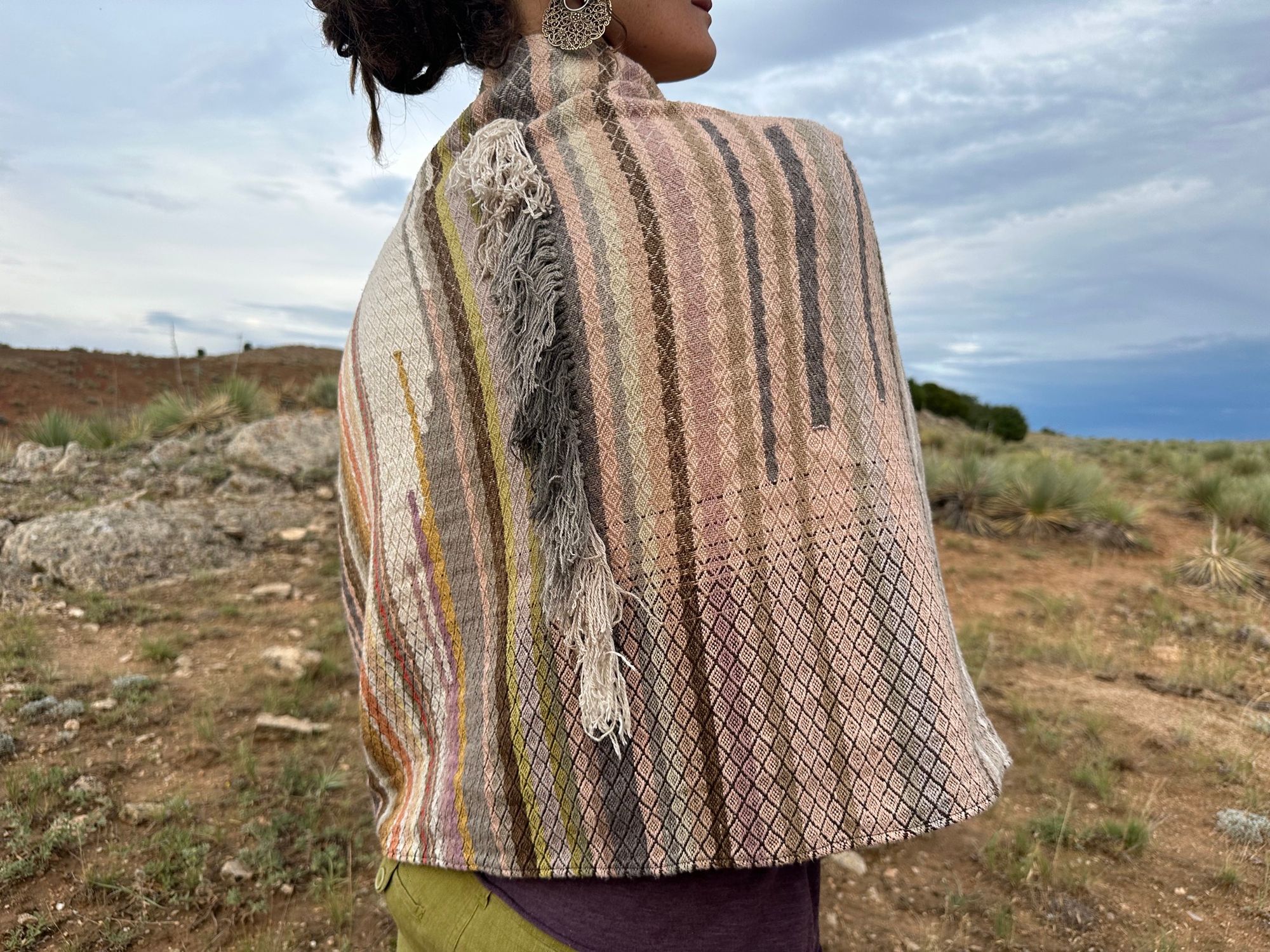 a woman wears a shawl of handwoven fabric of Naturally dyed subtle rainbow hues with a diamond texture pattern in a mountain landscape