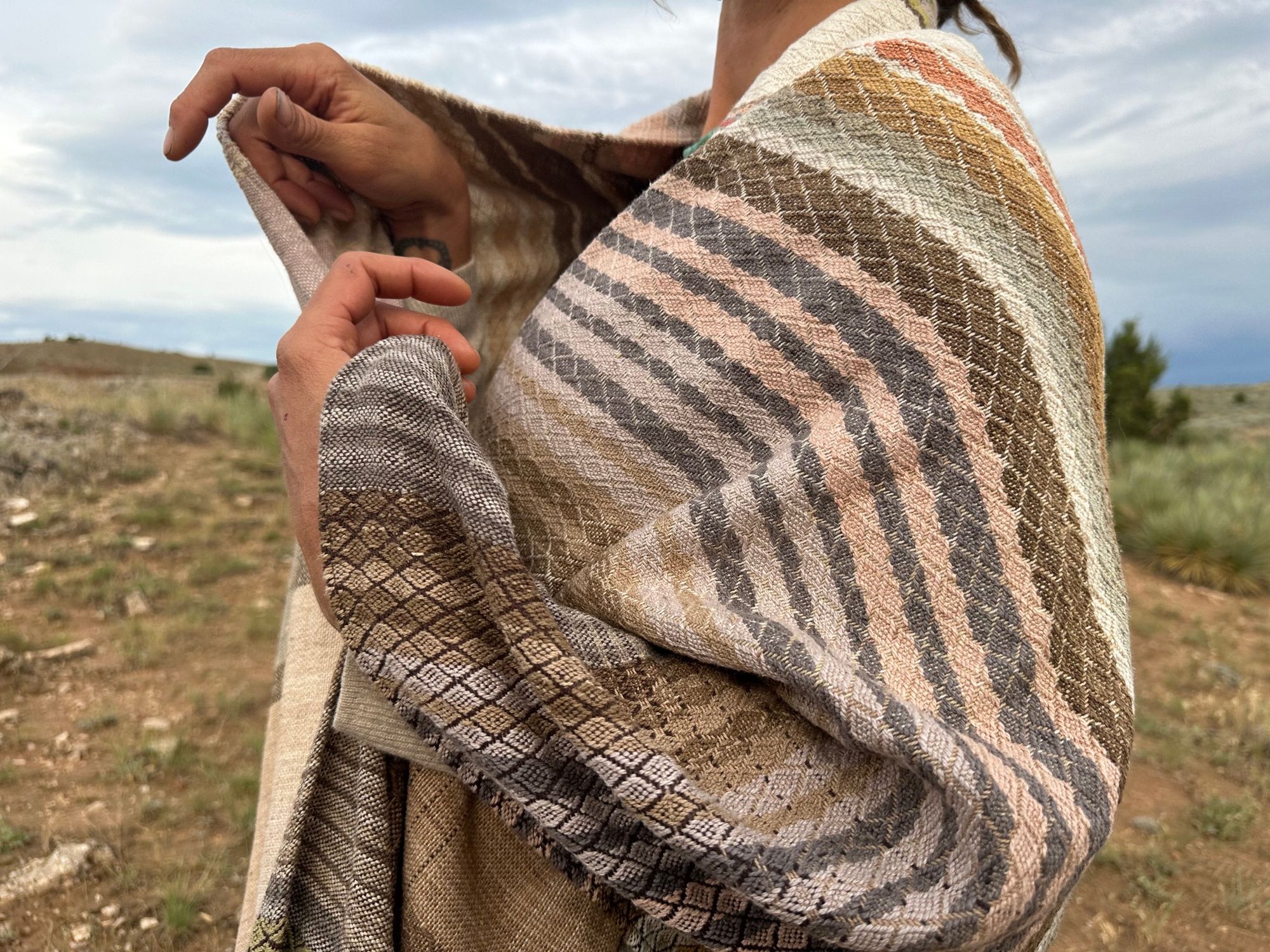 a woman wears a shawl of handwoven fabric of Naturally dyed subtle rainbow hues with a diamond texture pattern in a mountain landscape