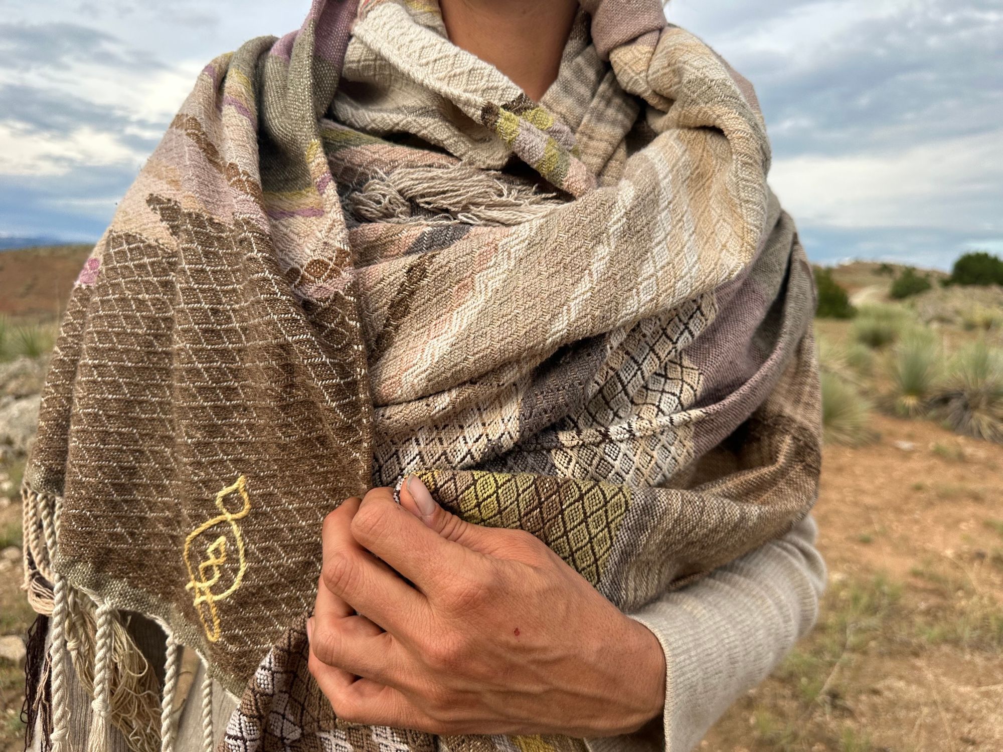 a woman wears a shawl of handwoven fabric of Naturally dyed subtle rainbow hues with a diamond texture pattern in a mountain landscape