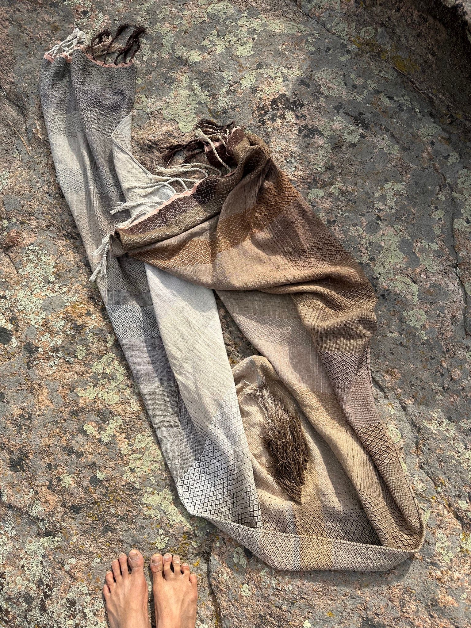 A detail of handwoven softly graded grey to white to brown diamond pattern raw silk fabric laying on a rock with barefeet in the foreground