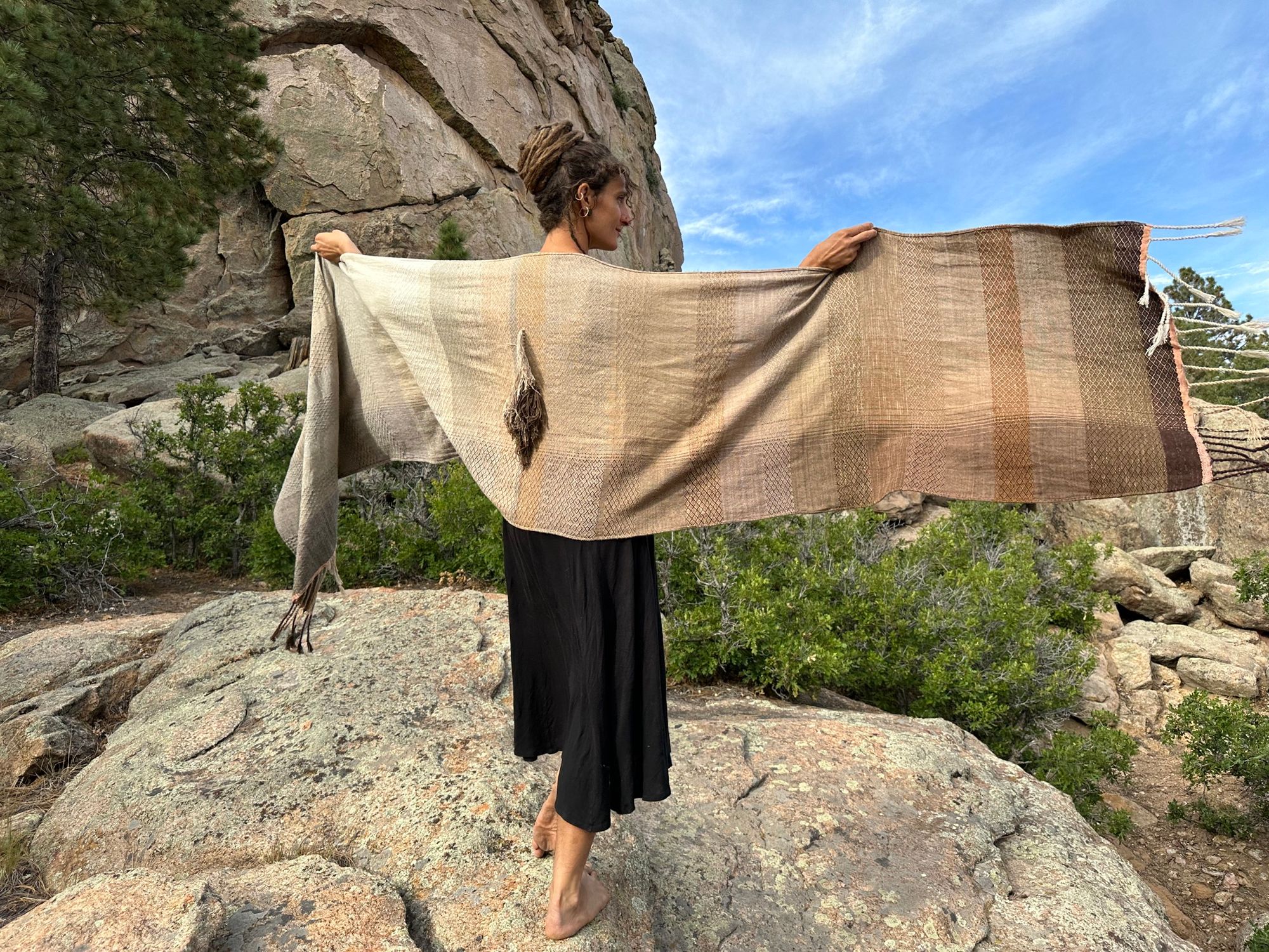 A detail of handwoven softly graded grey to white to brown diamond pattern raw silk fabricA woman in a black dress stands on large rocks wearing a handwoven naturally dyed grey, brown and white shawl