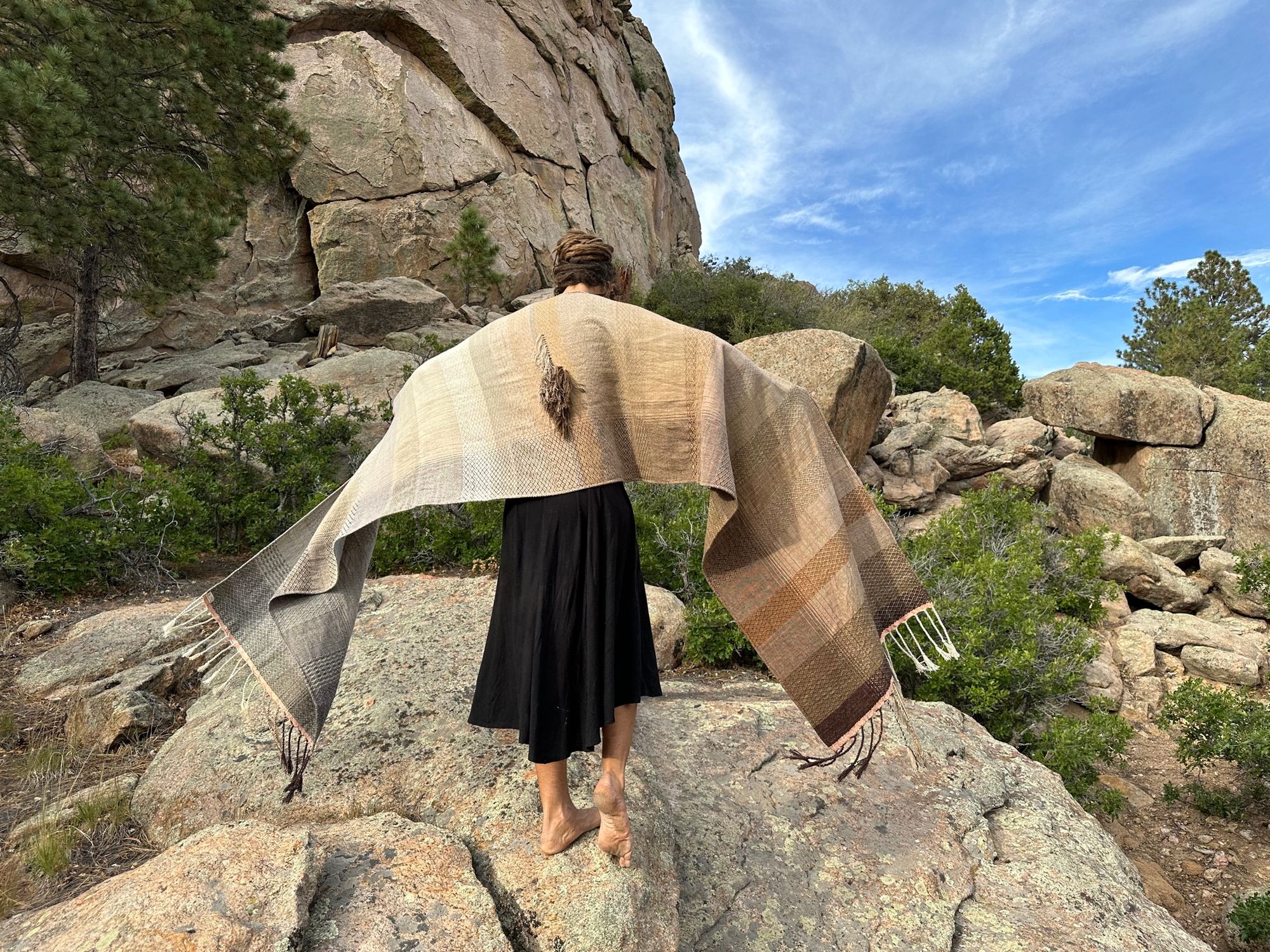 A woman in a black dress stands on large rocks wearing a handwoven naturally dyed grey, brown and white shawl