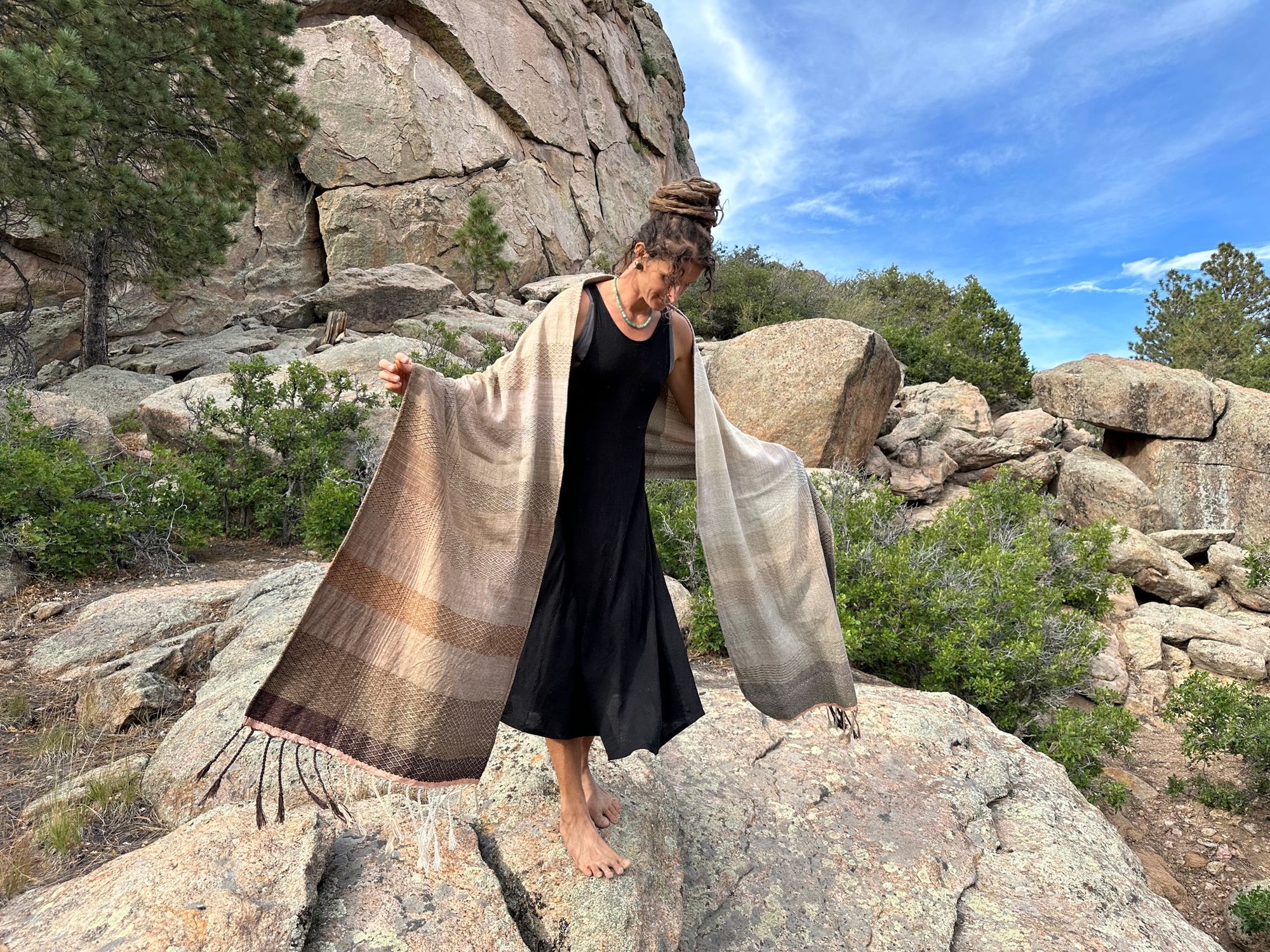 A woman in a black dress stands on large rocks wearing a handwoven naturally dyed grey, brown and white shawl