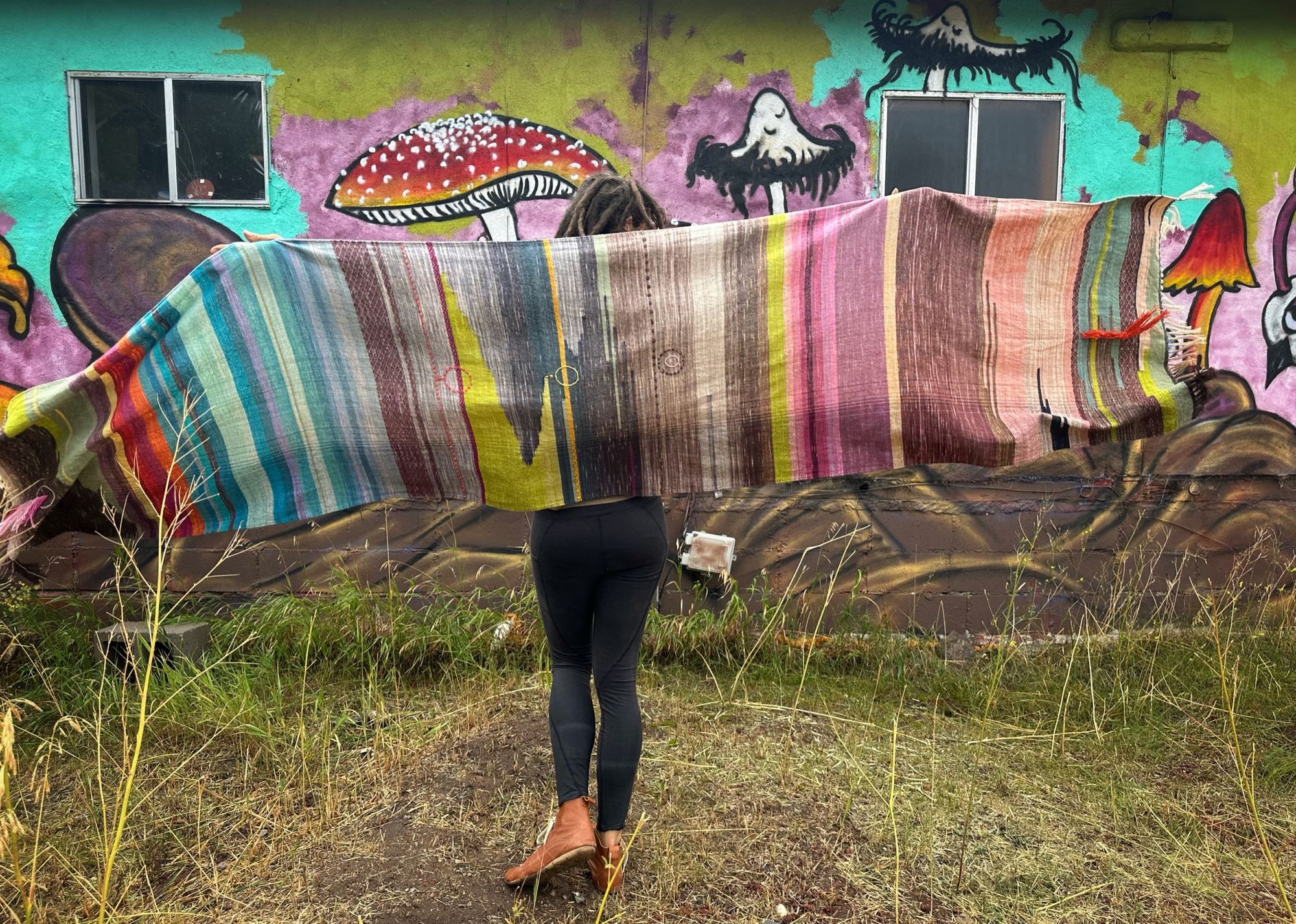 A woman wearing a raw silk handwoven fabric in a rainbow of hand dyed colors with circular moon-like details against a mural of mushrooms