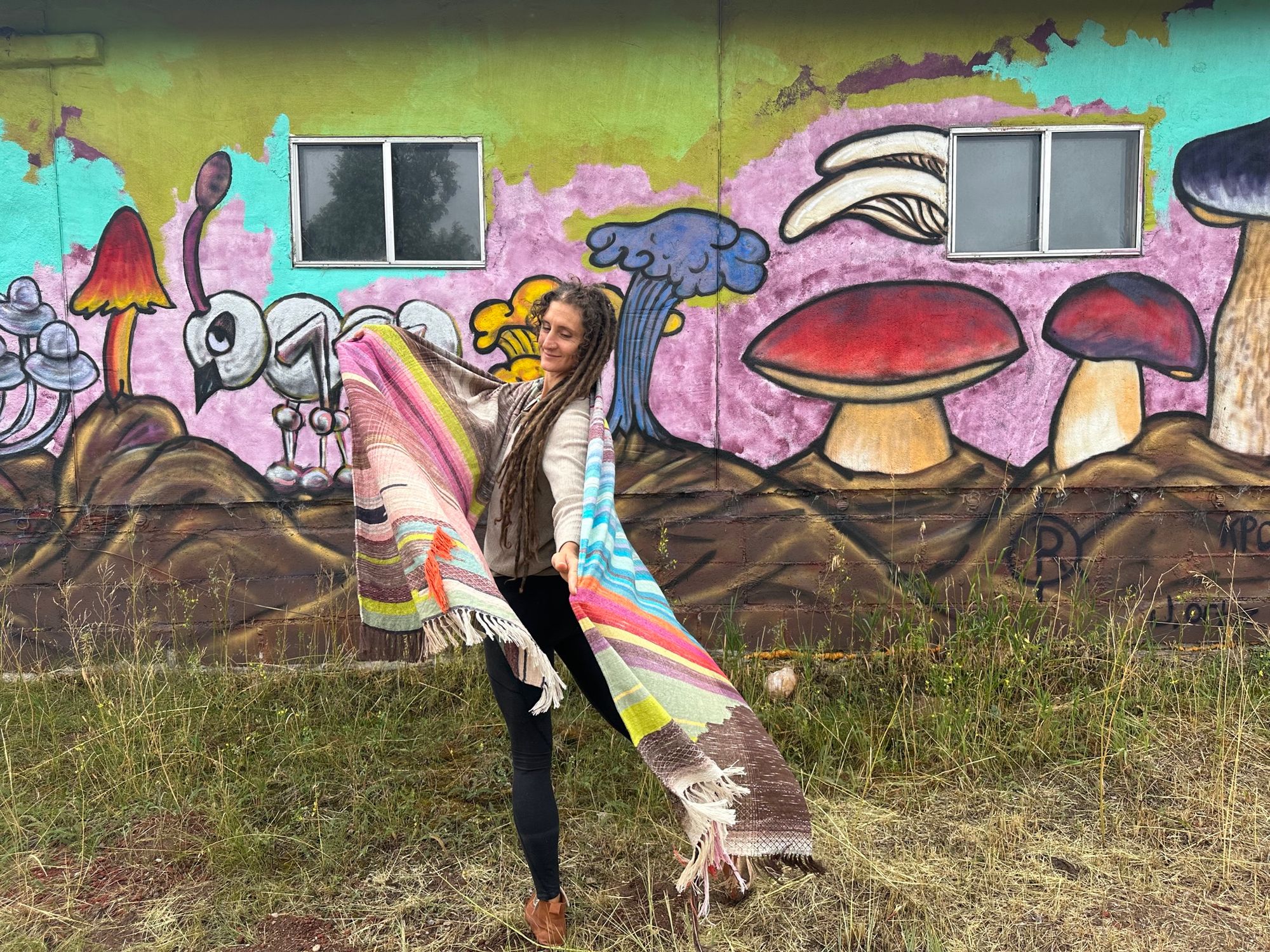 A woman wearing a raw silk handwoven fabric in a rainbow of hand dyed colors with circular moon-like details against a mural of mushrooms