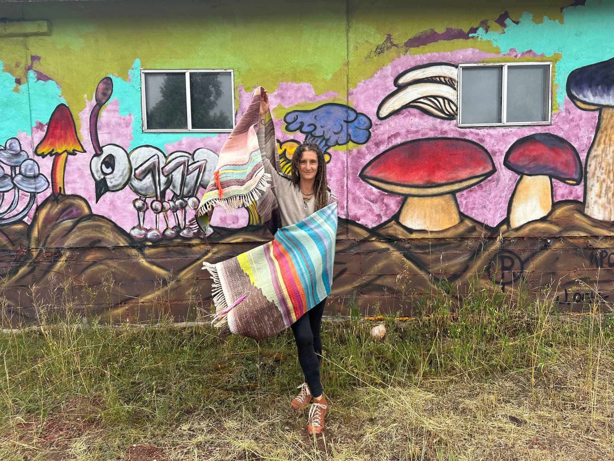 A woman wearing a raw silk handwoven fabric in a rainbow of hand dyed colors with circular moon-like details against a mural of mushrooms