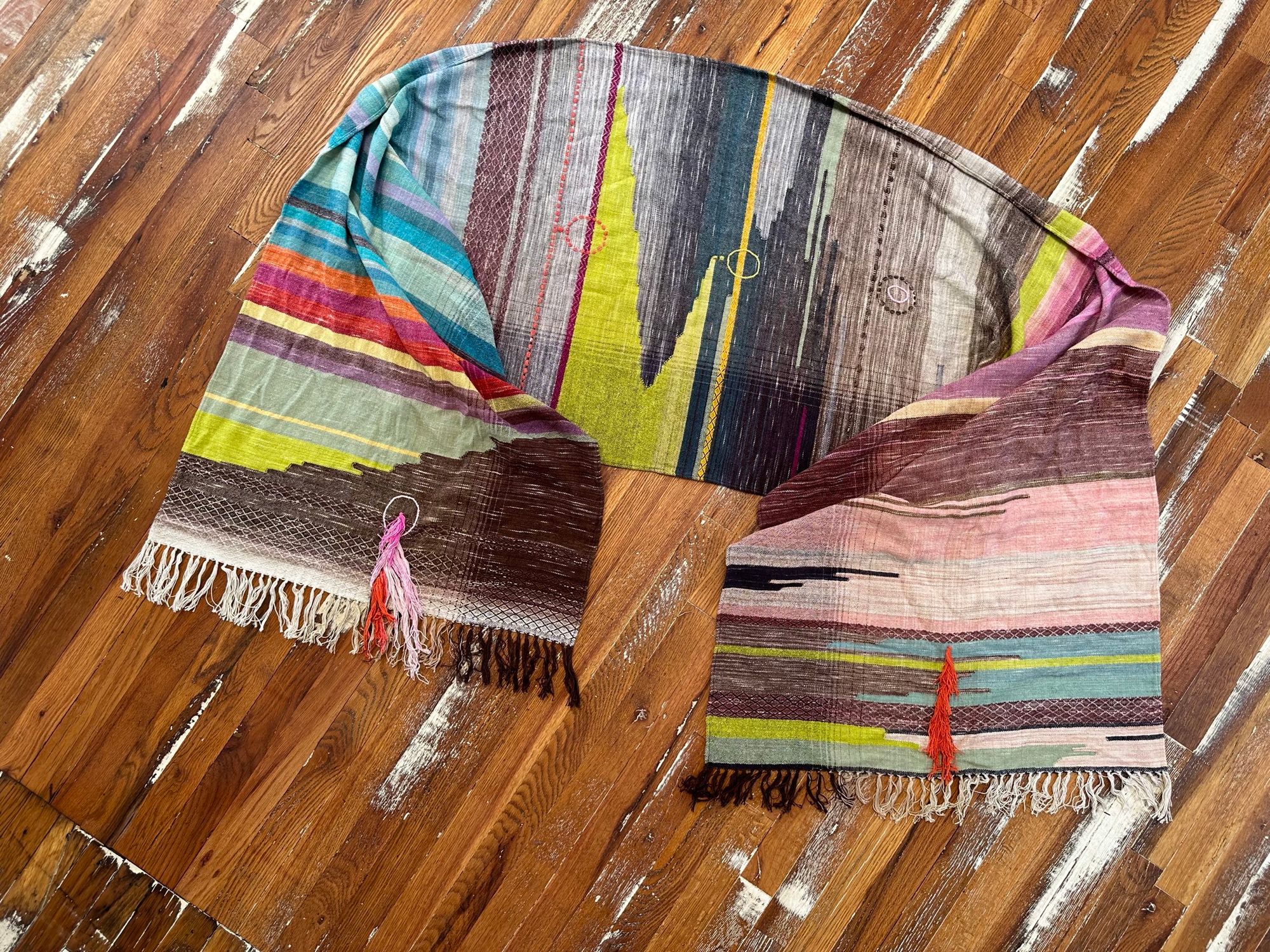 A detail of raw silk handwoven fabric in a rainbow of hand dyed colors with circular moon-like details laying on a wooden floor