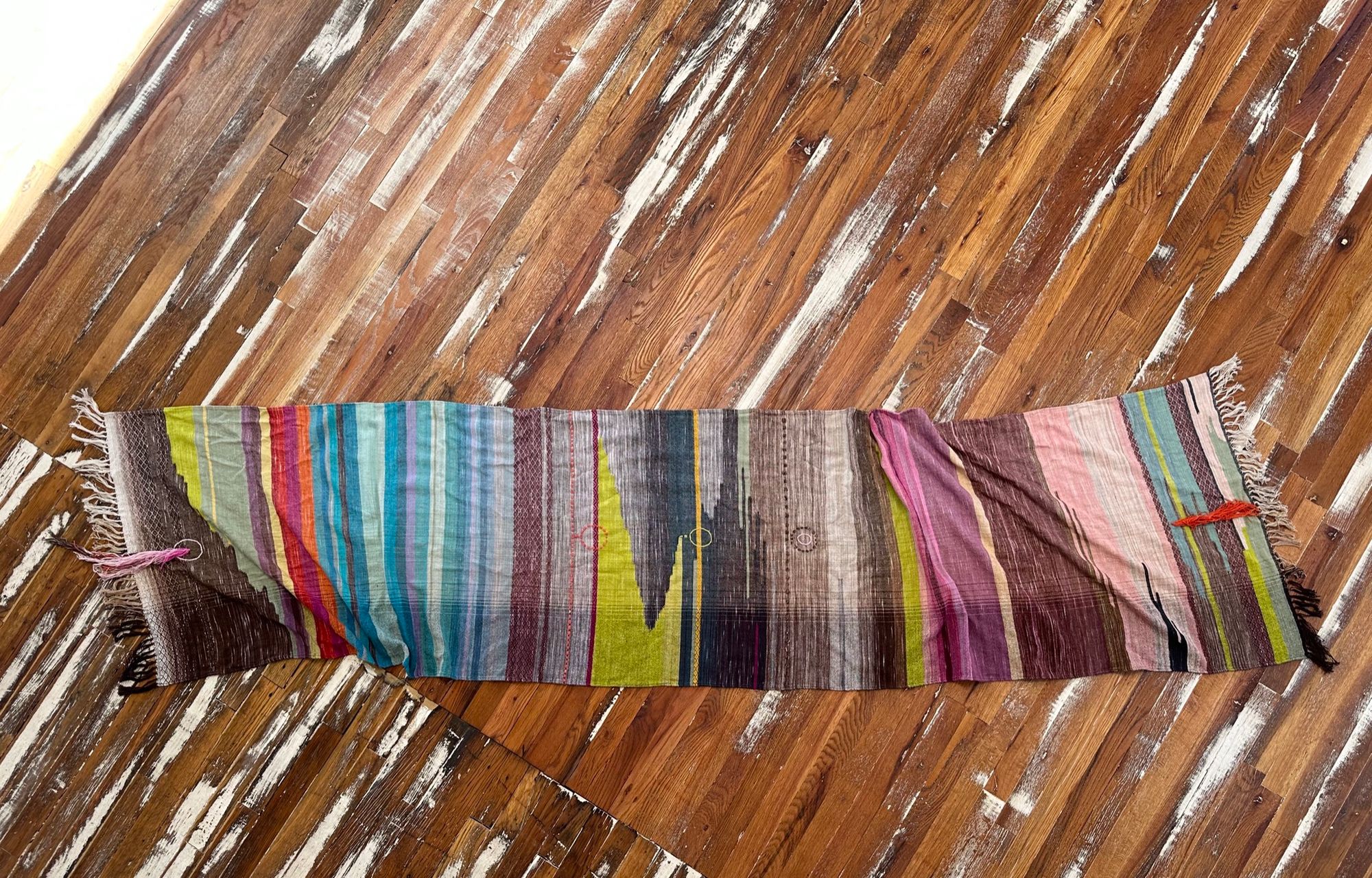 A detail of raw silk handwoven fabric in a rainbow of hand dyed colors with circular moon-like details laying on a wooden floor