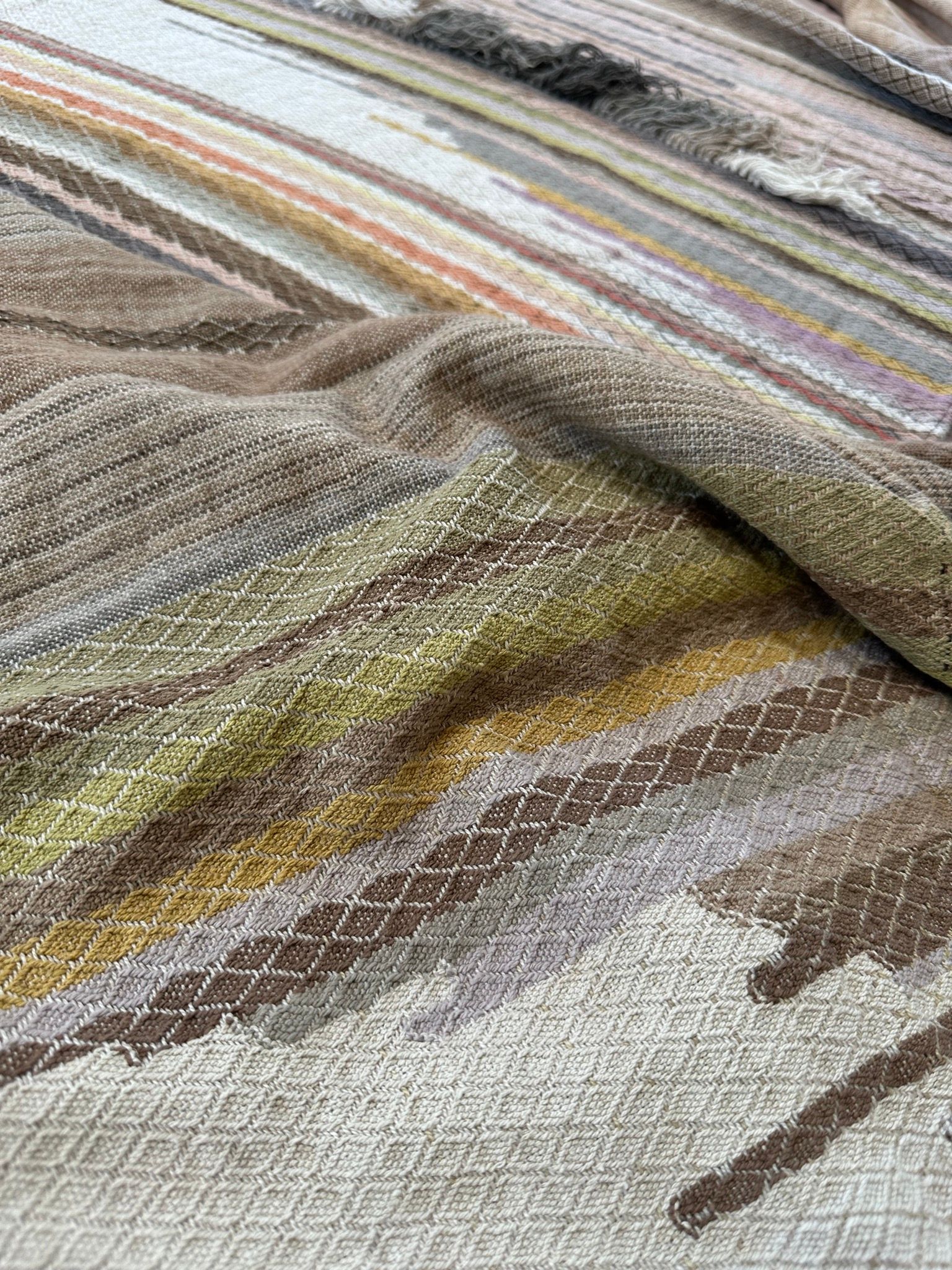 handwoven fabric of Naturally dyed subtle rainbow hues with a diamond texture pattern lays on a wooden floor