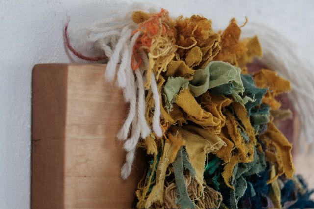 A fiber art sculpture made of yarn and scraps of fabric in a rainbow of colors with a clay face nestled into it, hanging from a maple wood frame on a white wall