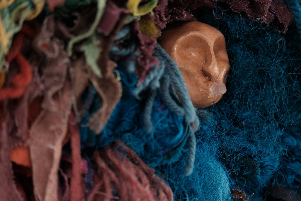 A fiber art sculpture made of yarn and scraps of fabric in a rainbow of colors with a clay face nestled into it, hanging from a maple wood frame on a white wall