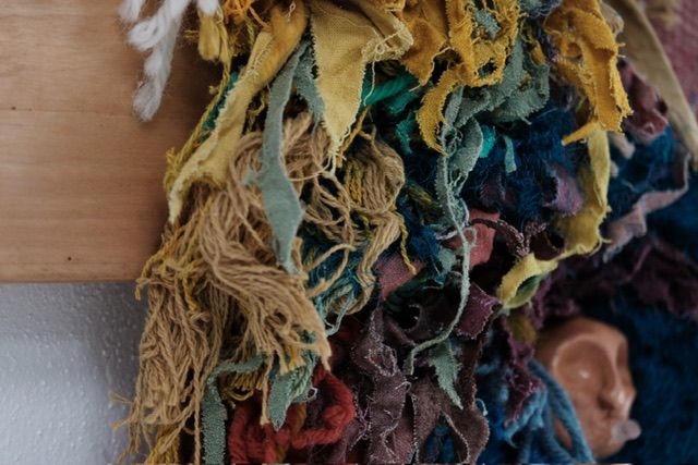 A fiber art sculpture made of yarn and scraps of fabric in a rainbow of colors with a clay face nestled into it, hanging from a maple wood frame on a white wall