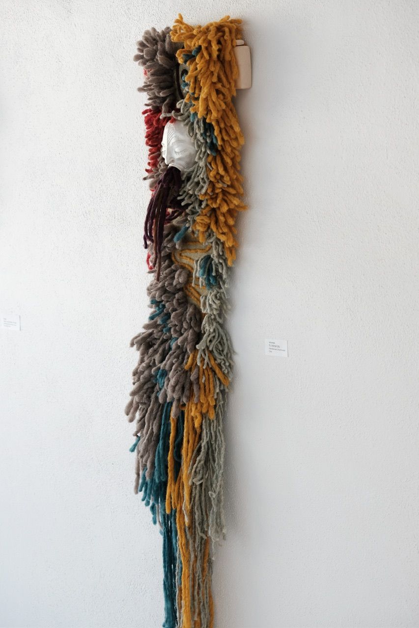 Handwoven many-colored pile woven churro wool wall hanging sculpture with a clay face emerging from it with purple yarn coming from its mouth
