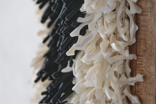 A handwoven sculptural necklace of black coral and mother of pearl hanging on a wooden frame On a white wall