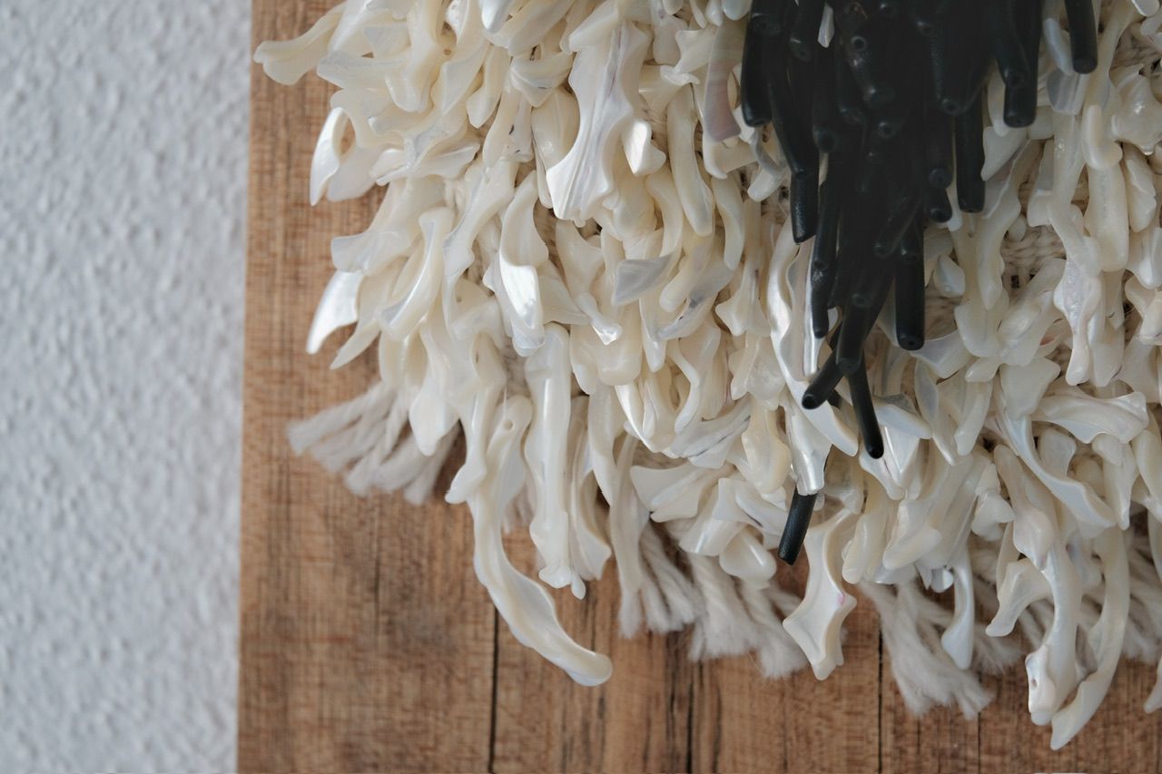 A handwoven sculptural necklace of black coral and mother of pearl hanging on a wooden frame On a white wall